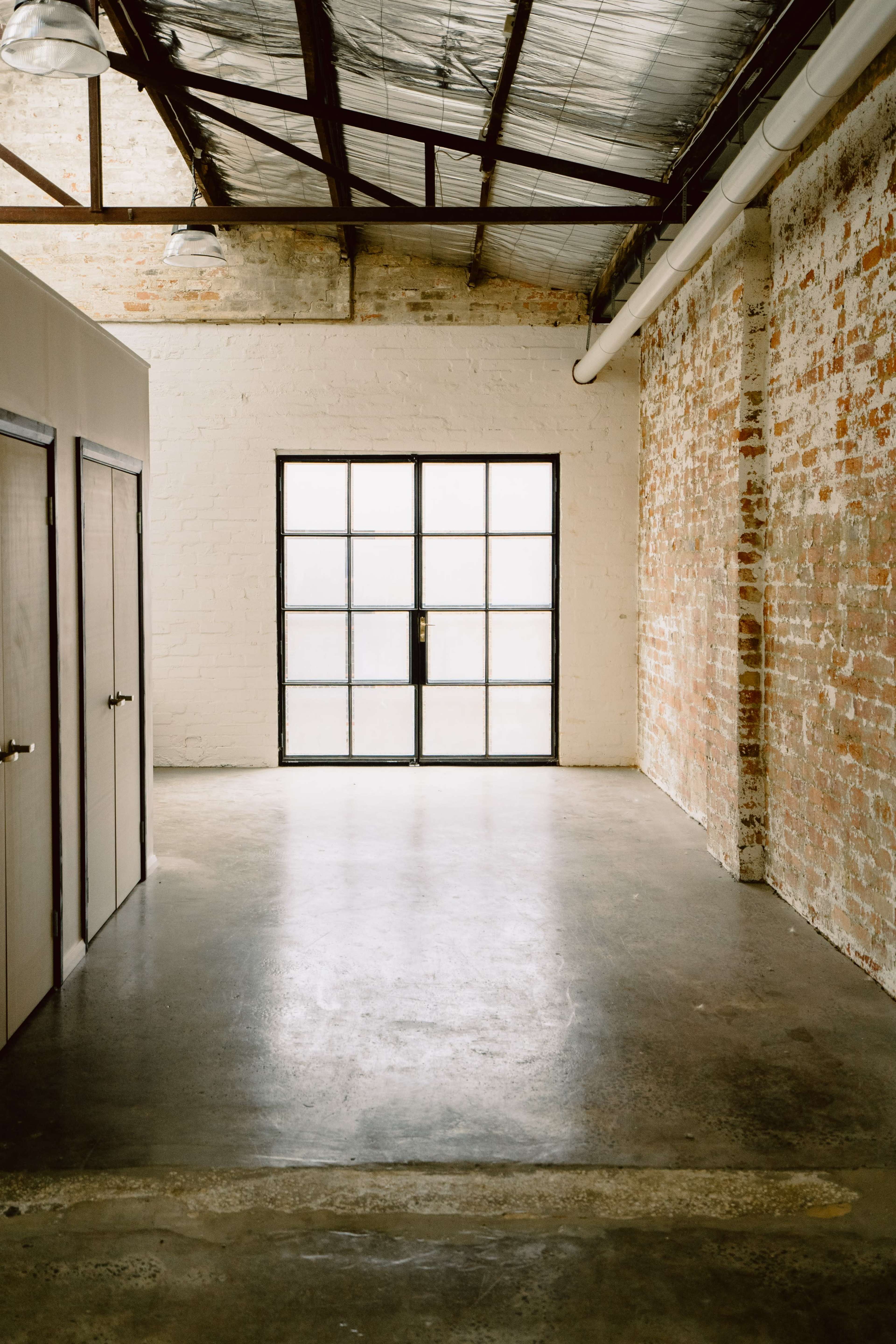 The image shows a spacious interior room with exposed brick walls, a concrete floor, and large glass doors leading to the outside.