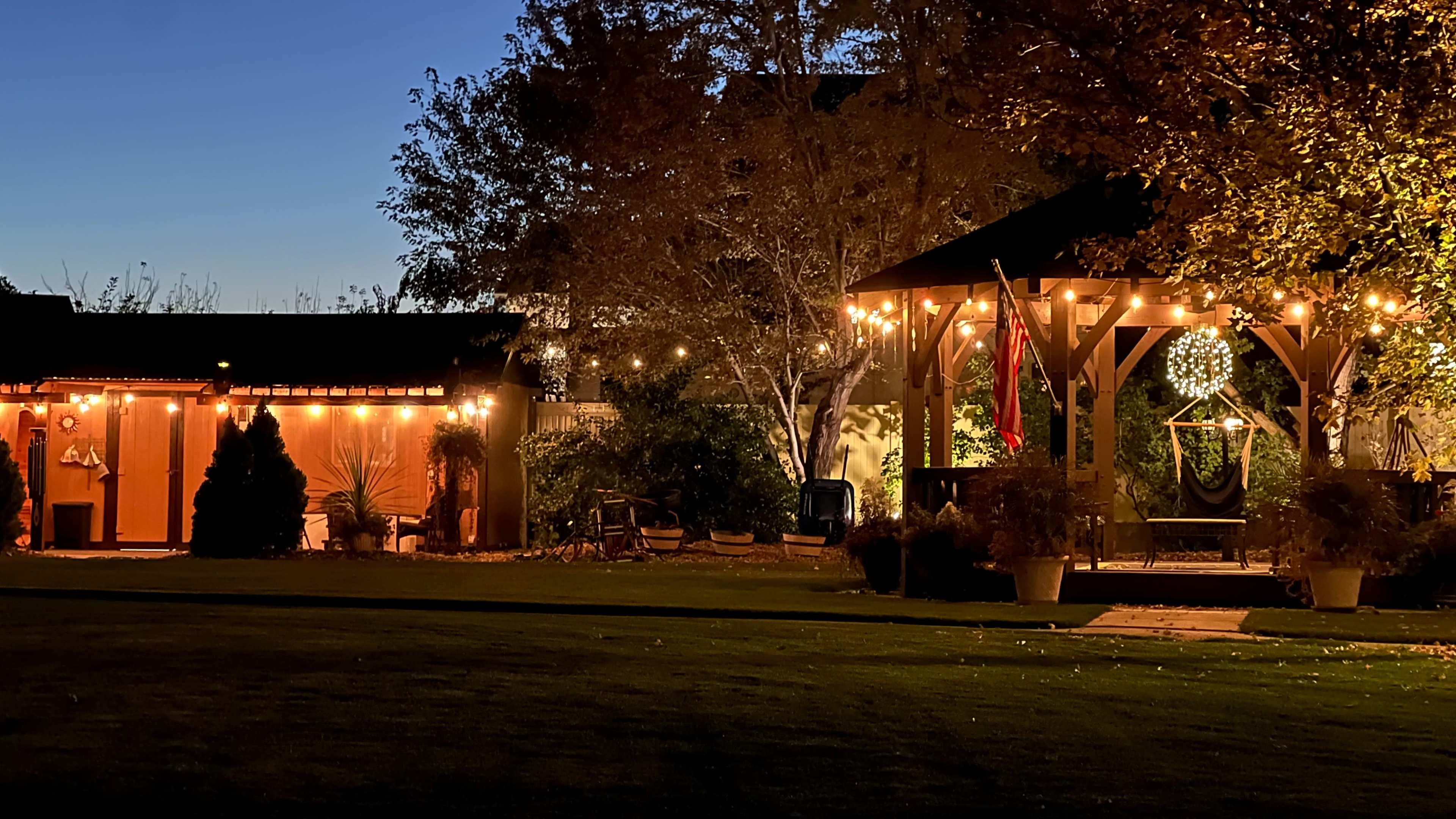 The image depicts a serene backyard scene at dusk, featuring a gazebo adorned with string lights and a cozy seating area, surrounded by well-maintained greenery.