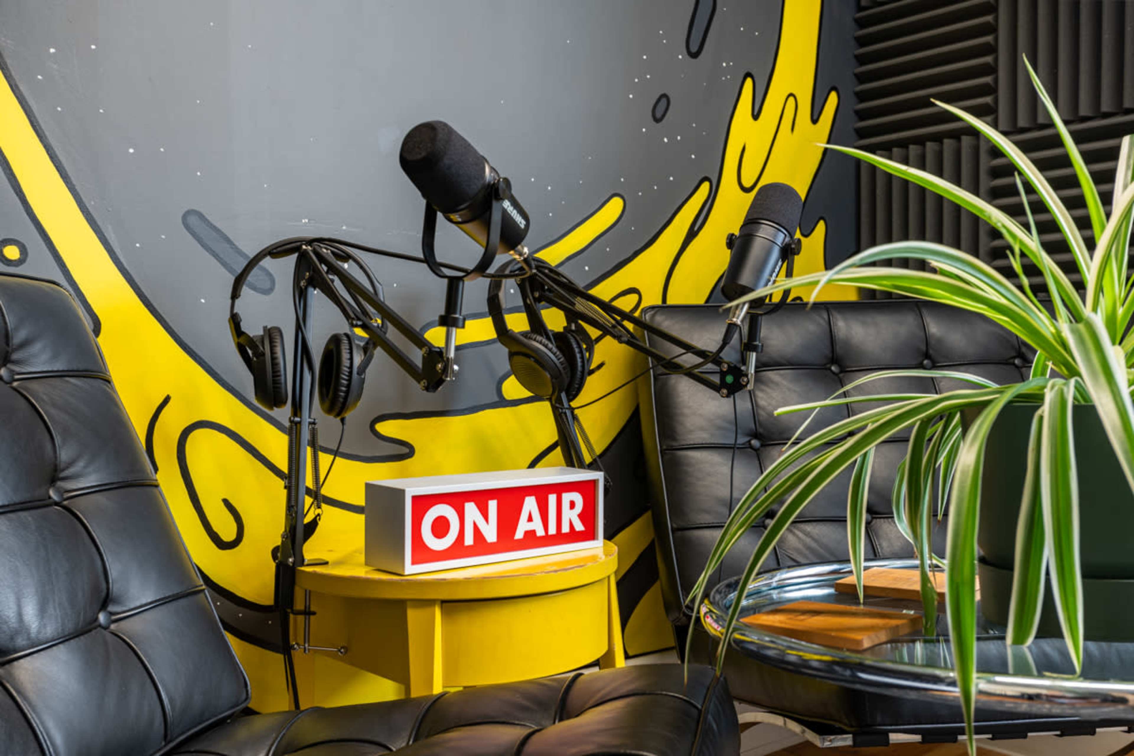 A recording studio setup with two black chairs, a "ON AIR" sign on a yellow table, and two microphones on adjustable arms beside headphones.