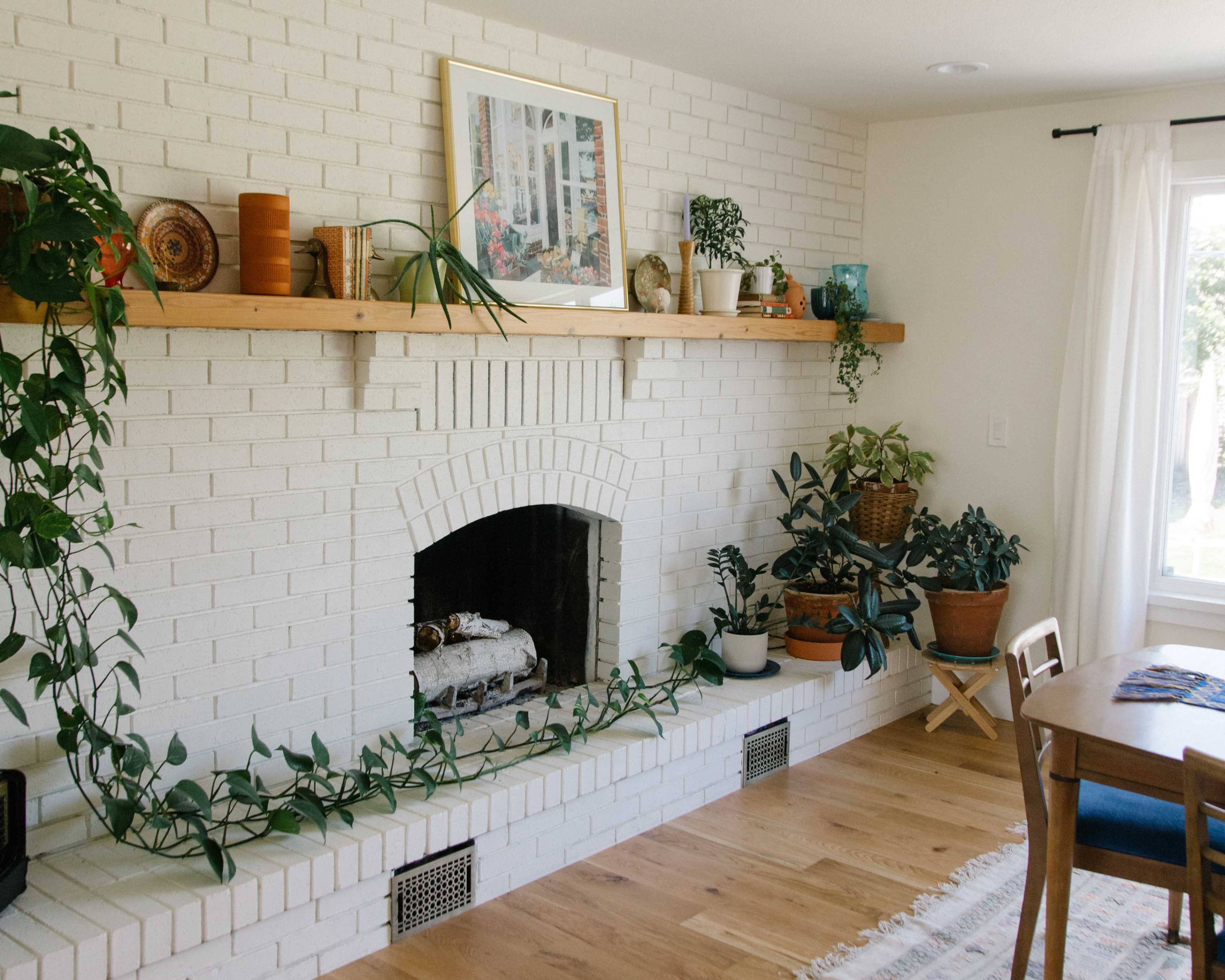 A bright living room features a white brick fireplace with a wooden shelf above, adorned with plants and decorative items.