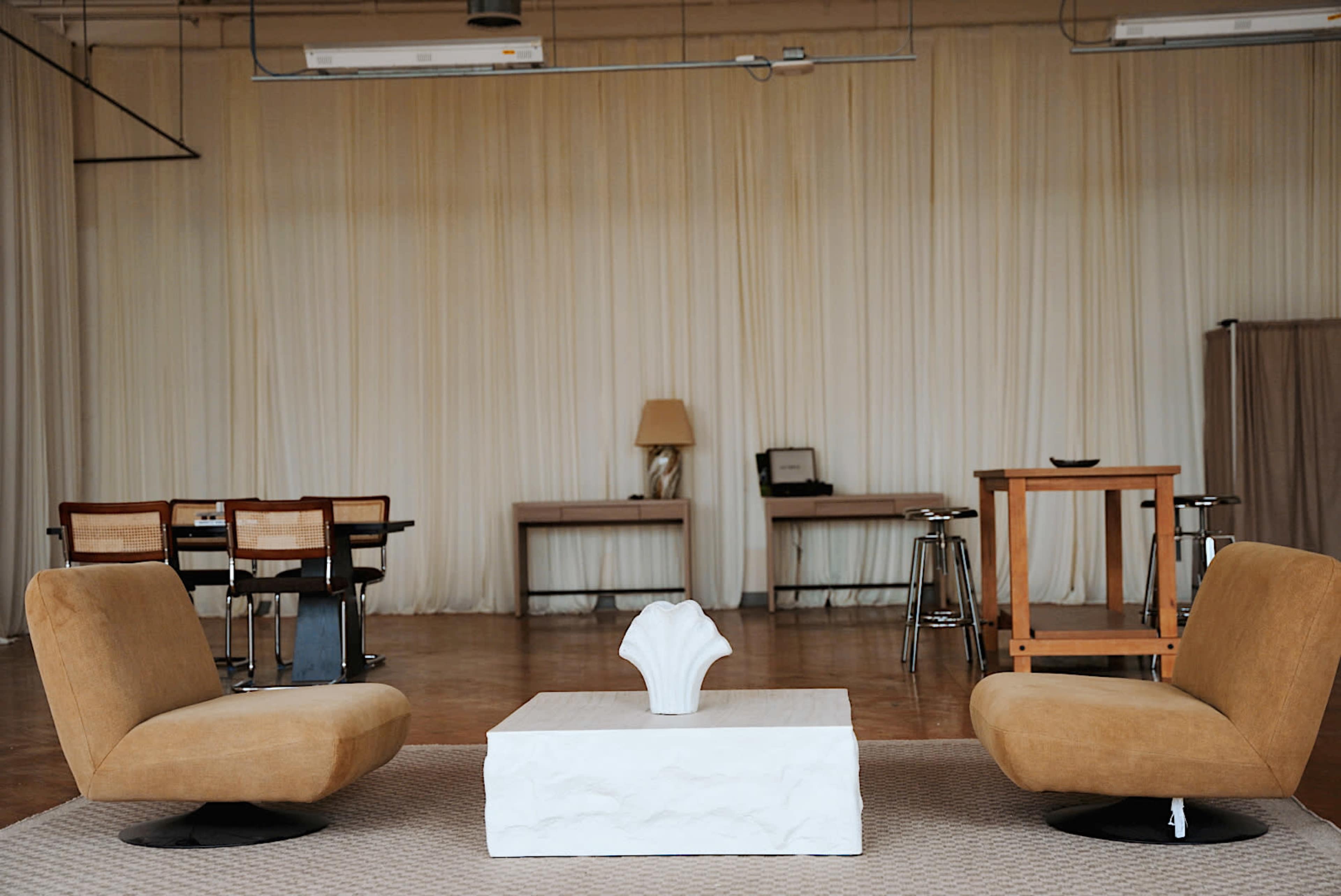 The image shows a minimalist seating area with two tan chairs flanking a white coffee table, set against a backdrop of draped fabric and two wooden desks in the background.