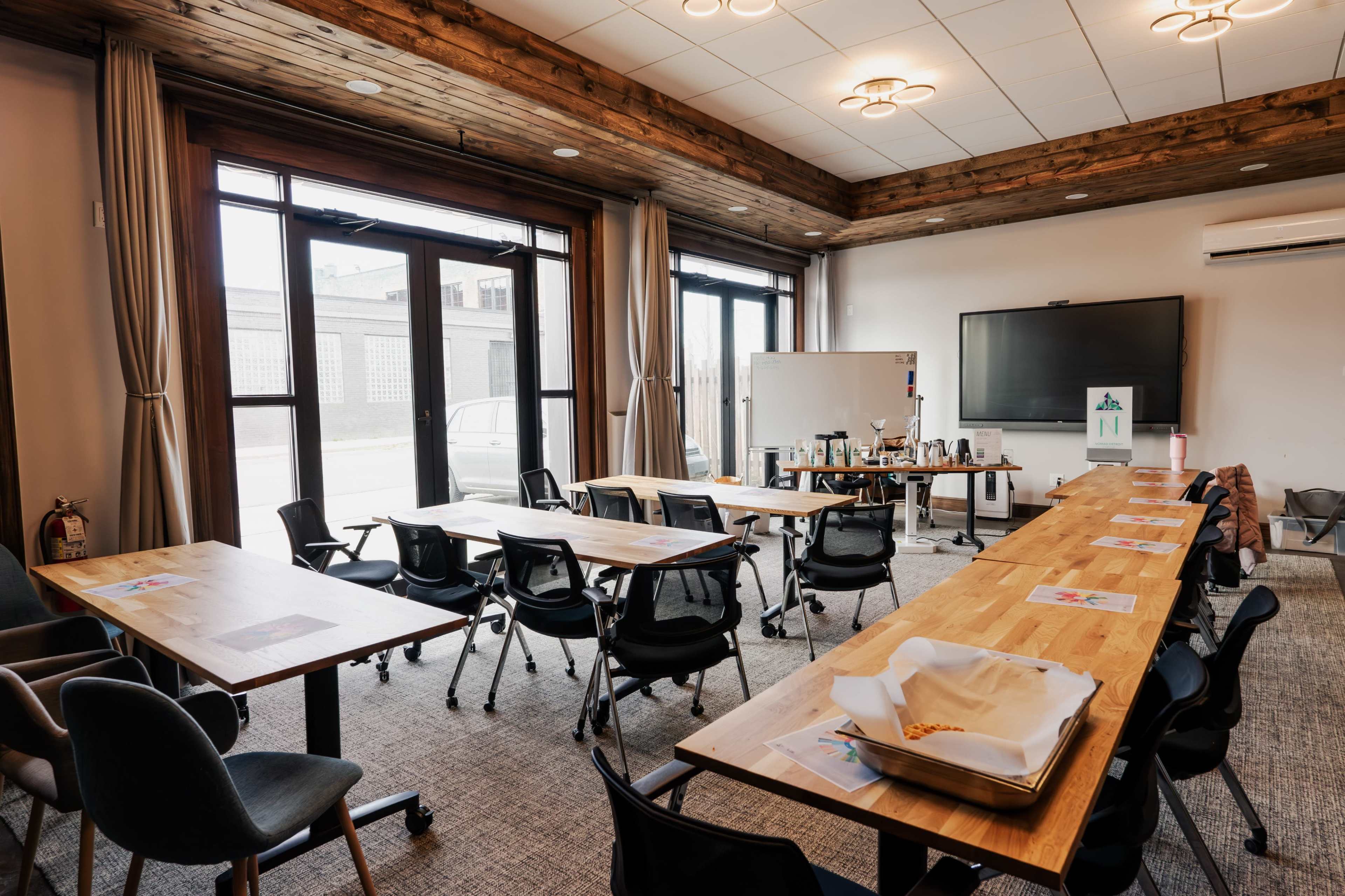 A conference room features several wooden tables arranged with chairs, a large screen, and refreshments in the background.