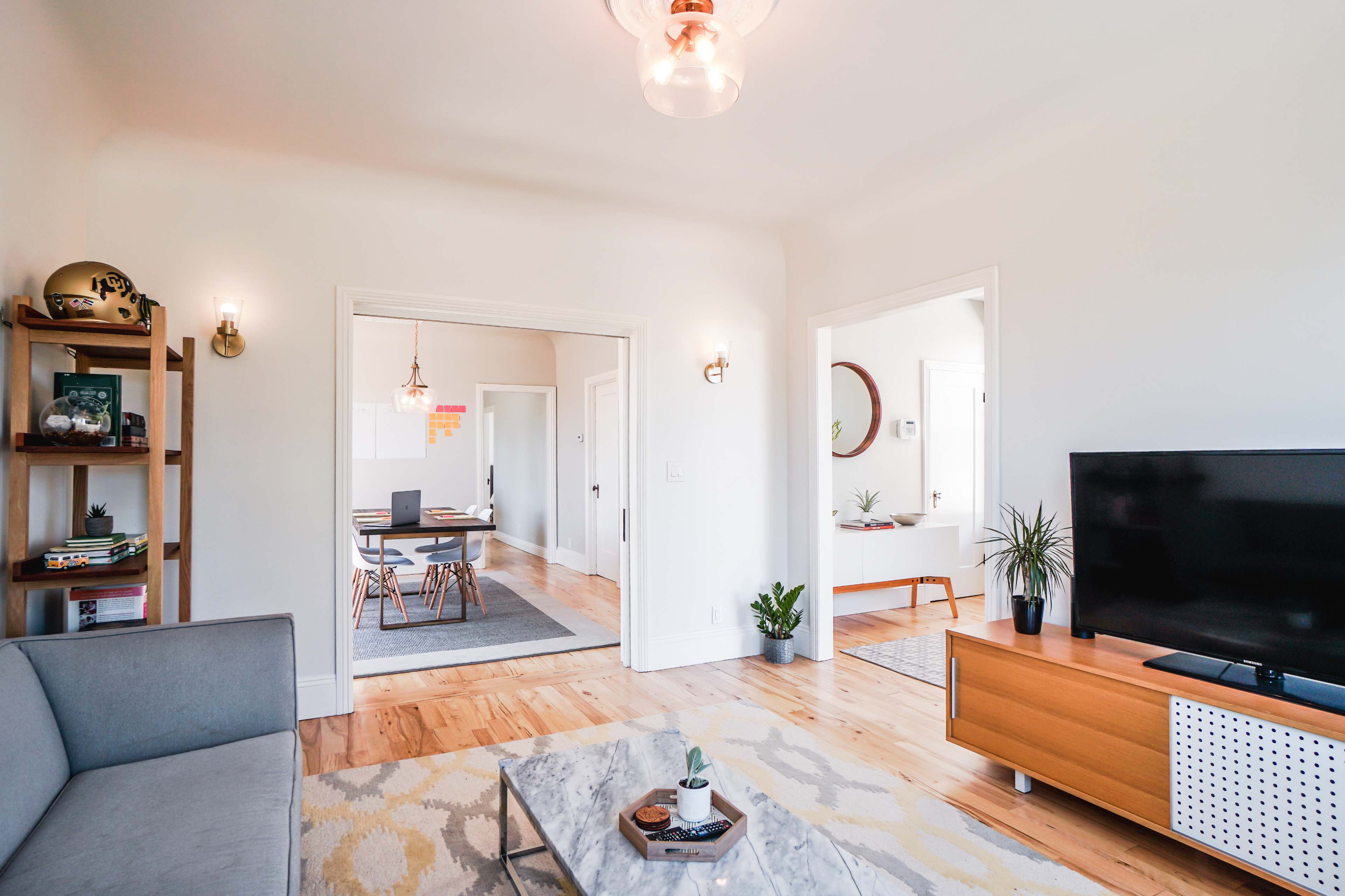 A modern living room features a gray sofa, a coffee table with a plant, and a large television, with an adjacent dining area visible through an open doorway.