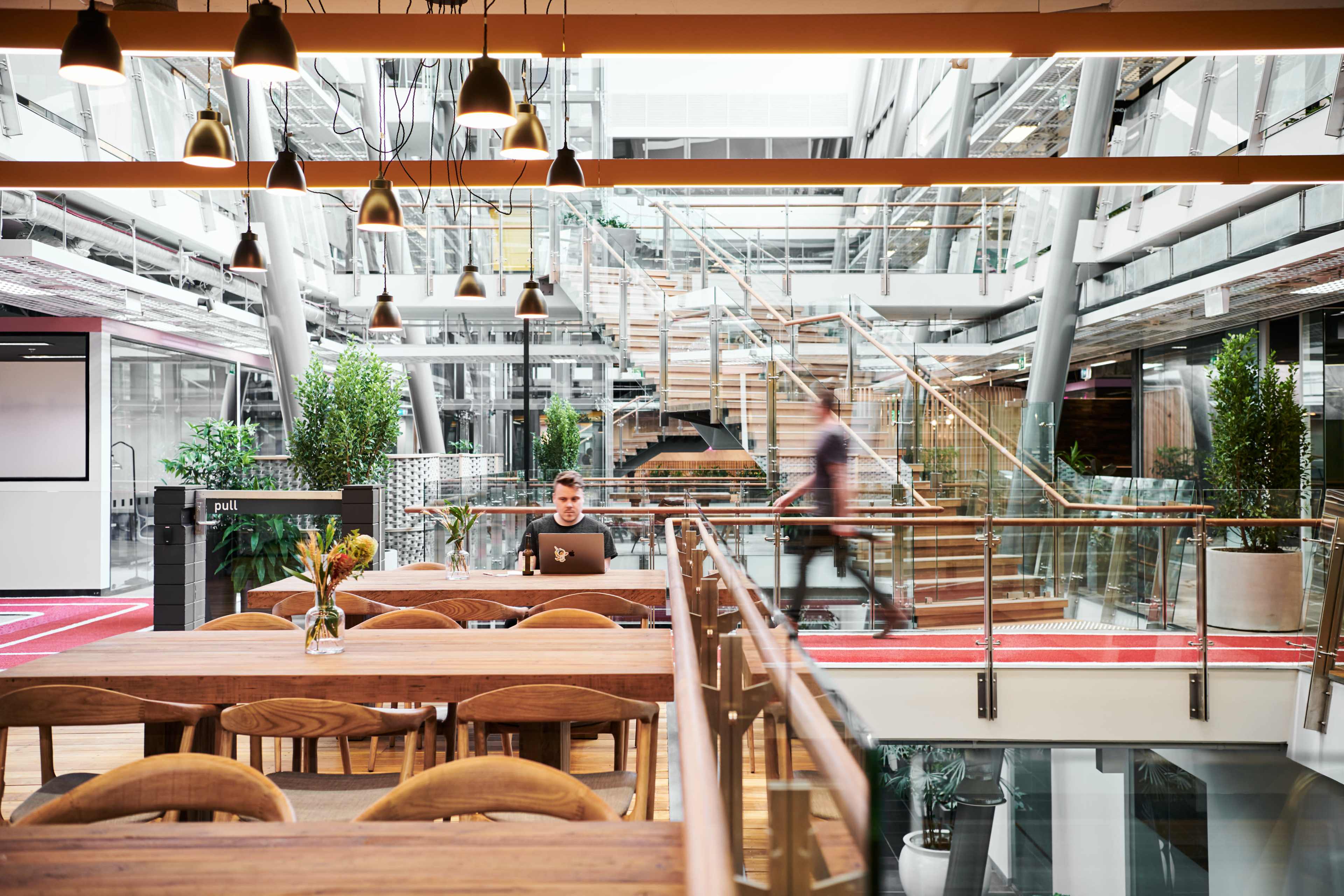 The image shows a modern open-plan office with wooden tables, plants, and a person working on a laptop, while a blurred individual walks in the background.