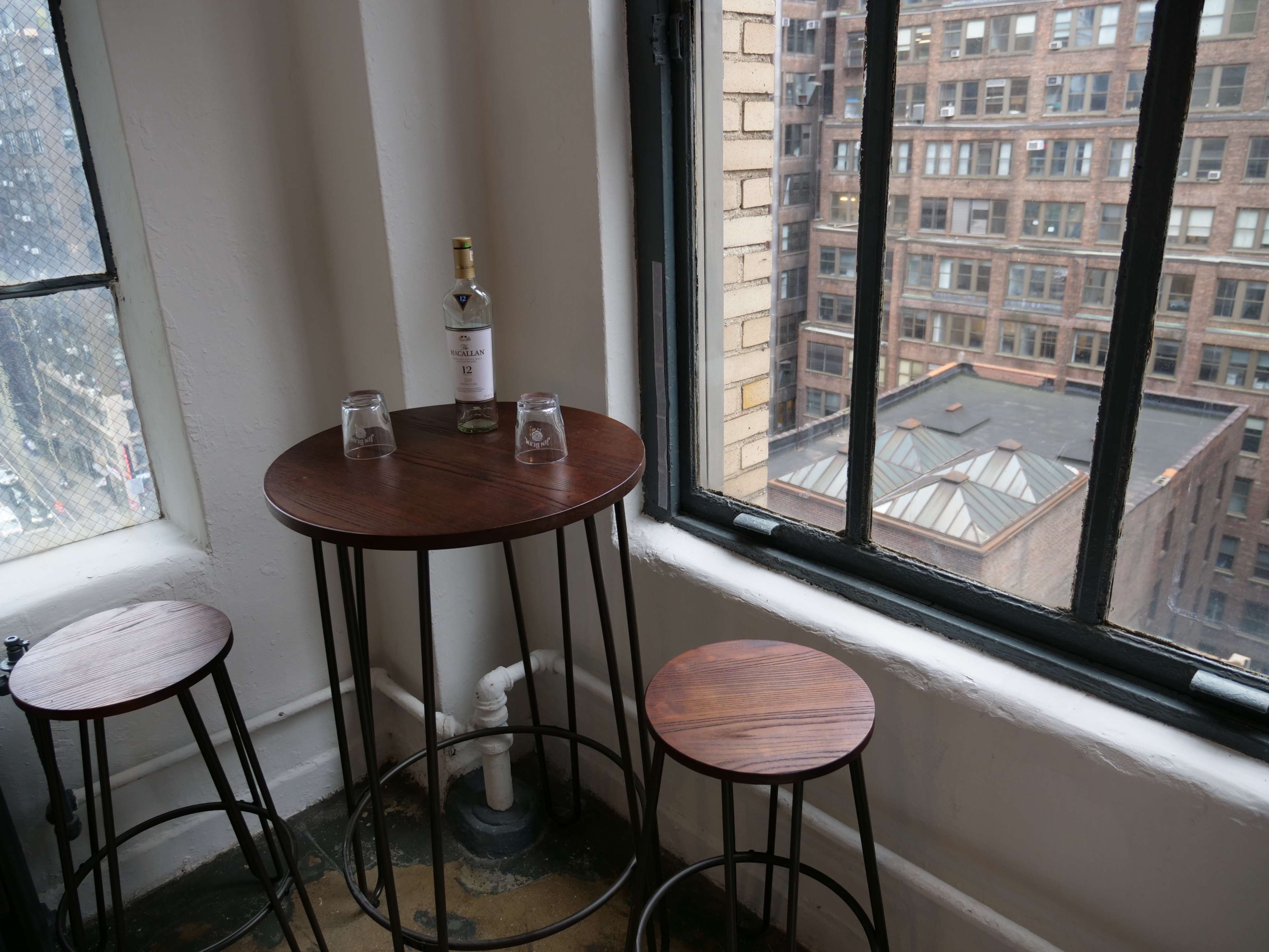 A small wooden table with two stools and a bottle of liquor sits near a large window overlooking a cityscape.