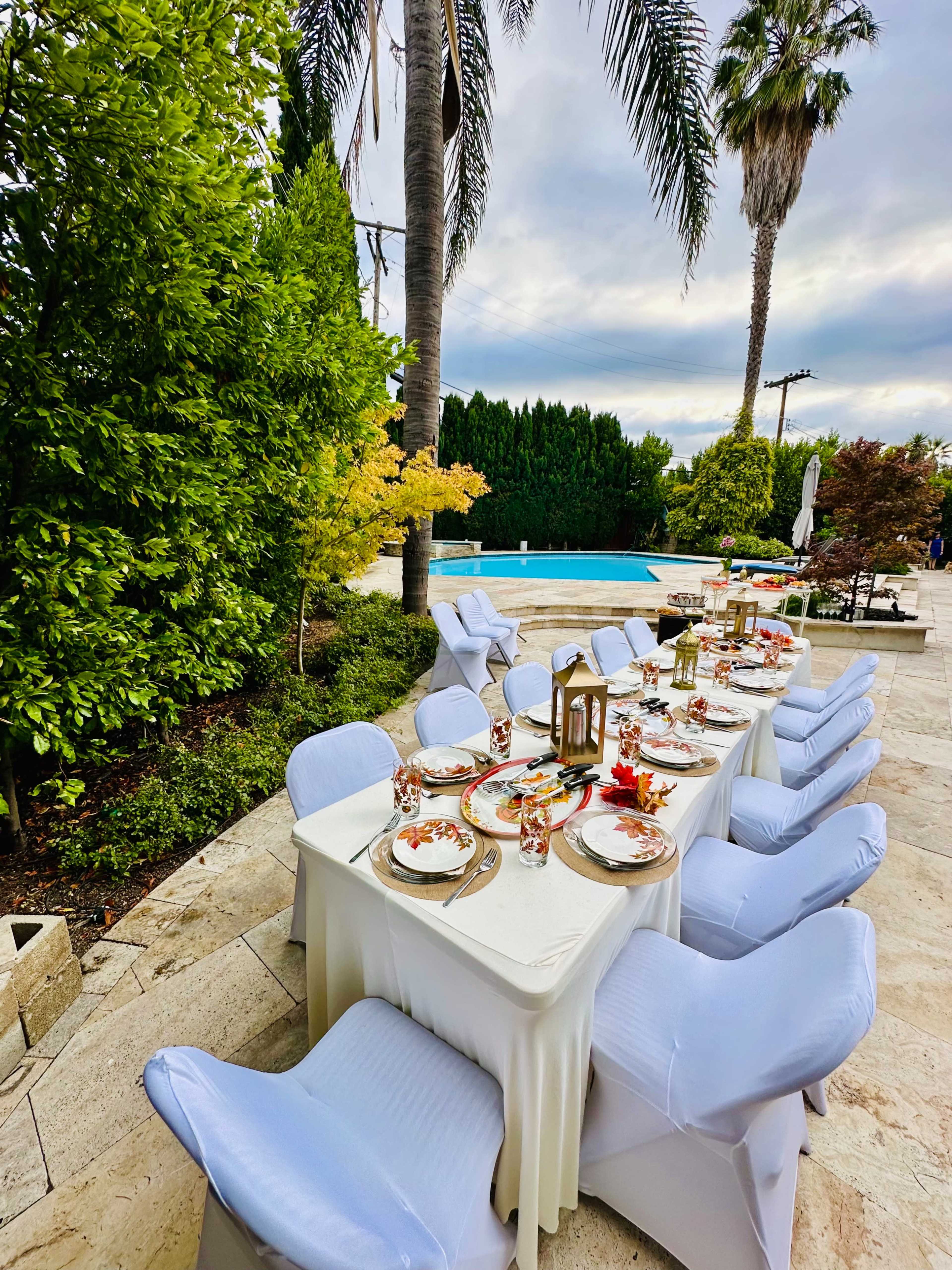 A long dining table is set with plates and decor, surrounded by chairs, near a swimming pool and lush greenery.
