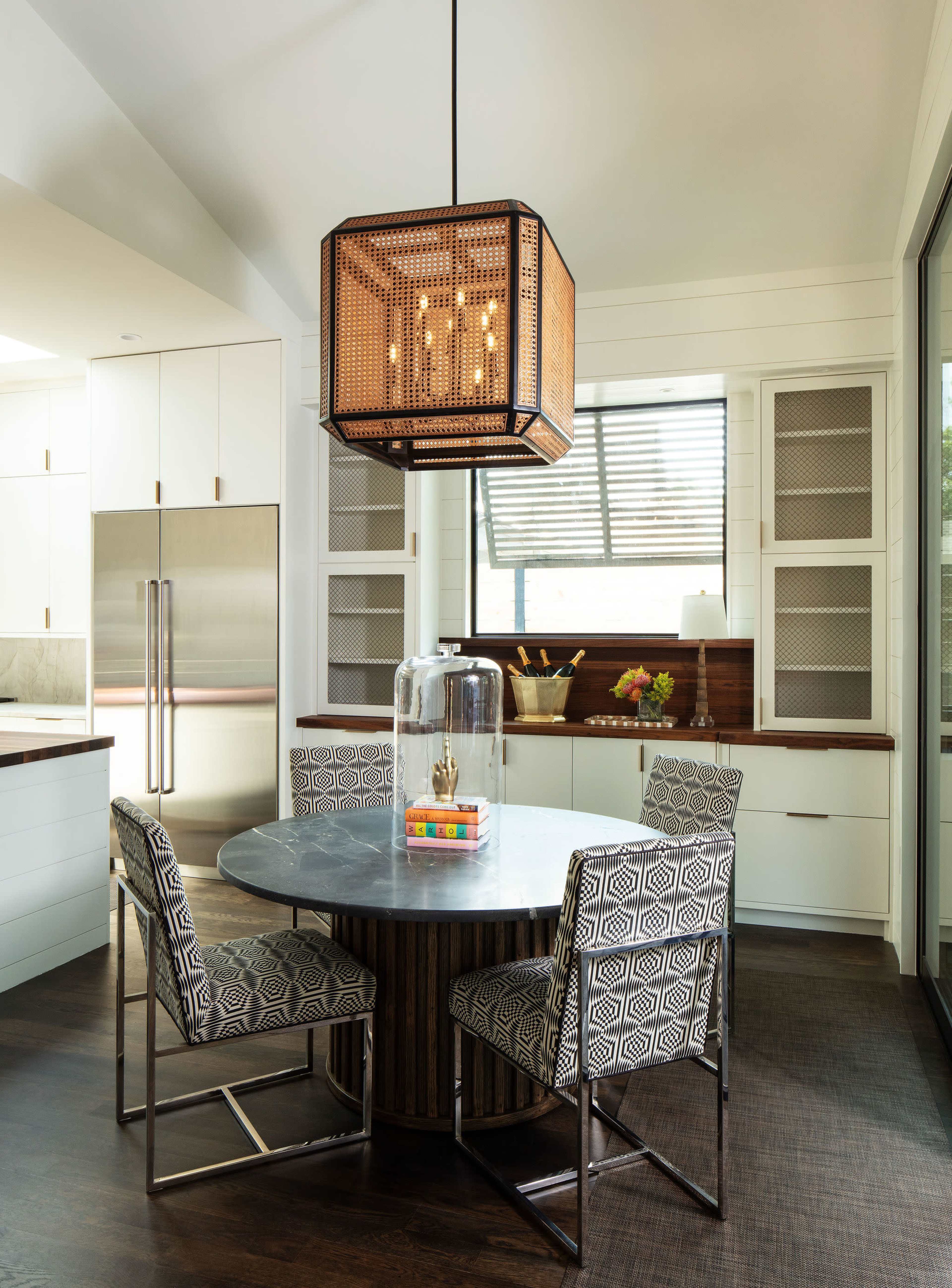 The image shows a modern kitchen featuring a round dining table surrounded by patterned chairs, beneath a large geometric pendant light.
