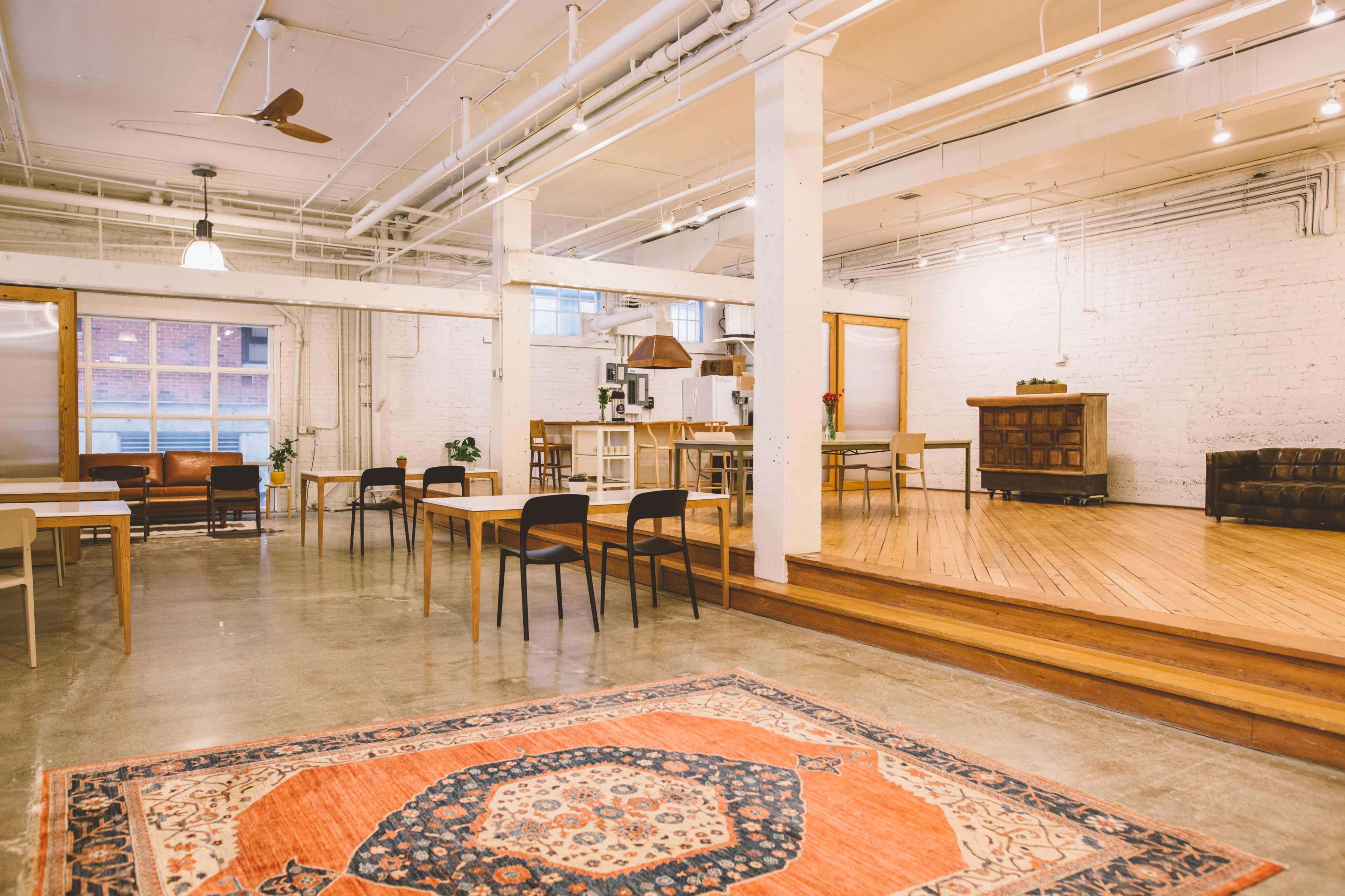 A spacious, modern indoor environment with wooden floors, white brick walls, tables with chairs, and a cozy area featuring a sofa and a rug.