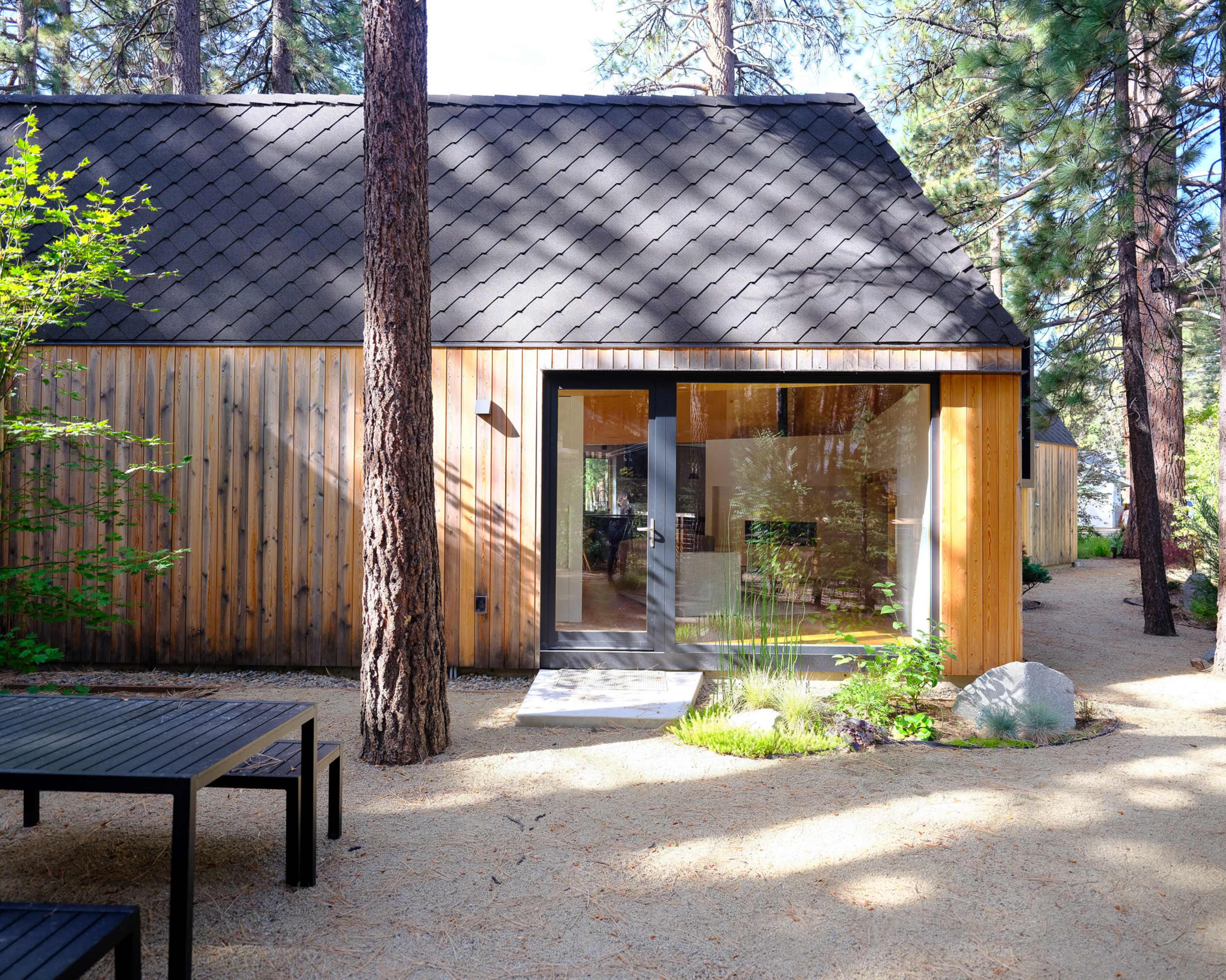 The image shows a modern wooden cabin with large glass doors set among tall trees and a gravel pathway.