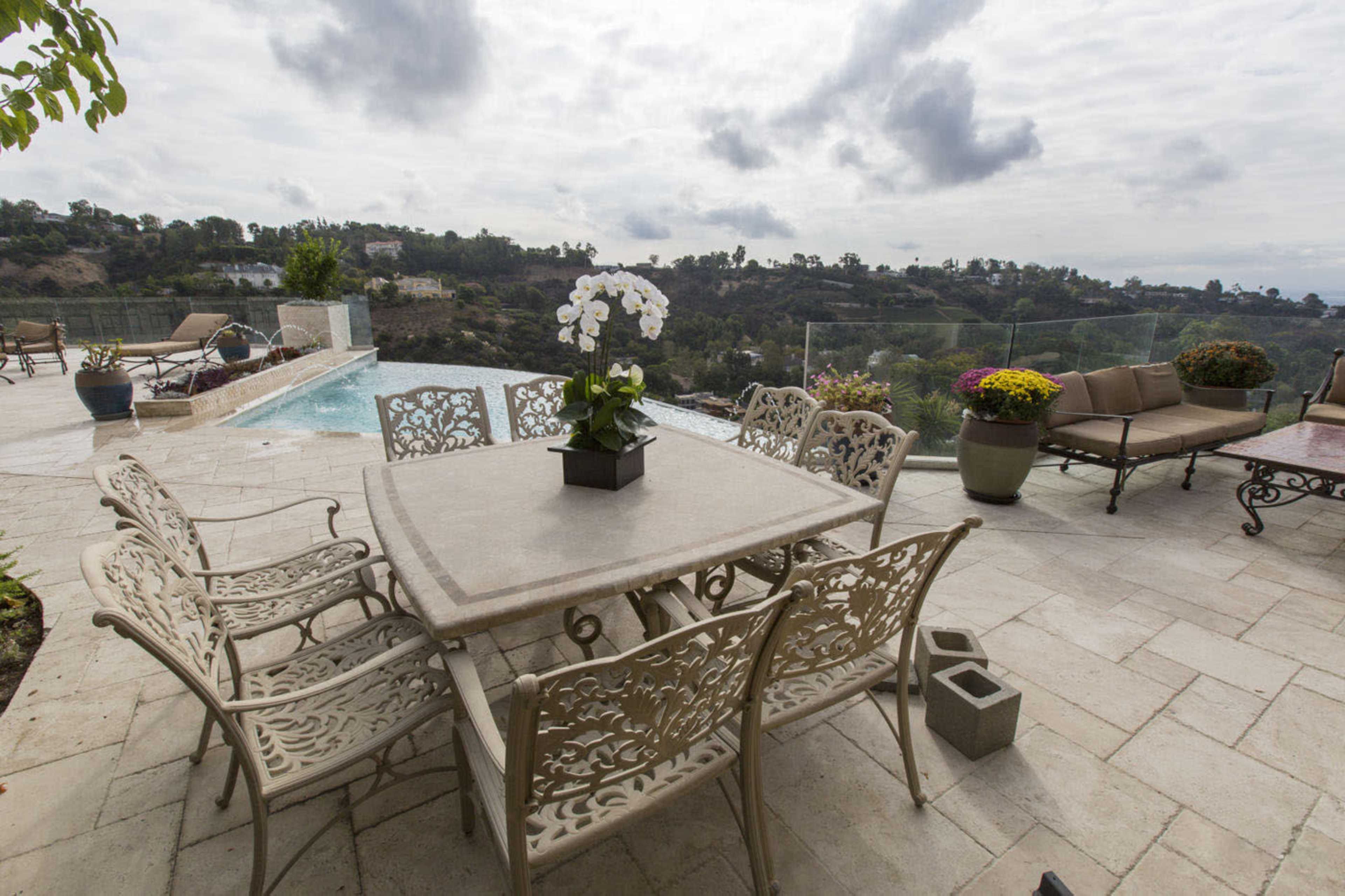 A rectangular patio table surrounded by eight chairs sits beside a pool with scenic views of hills and clouds in the background.