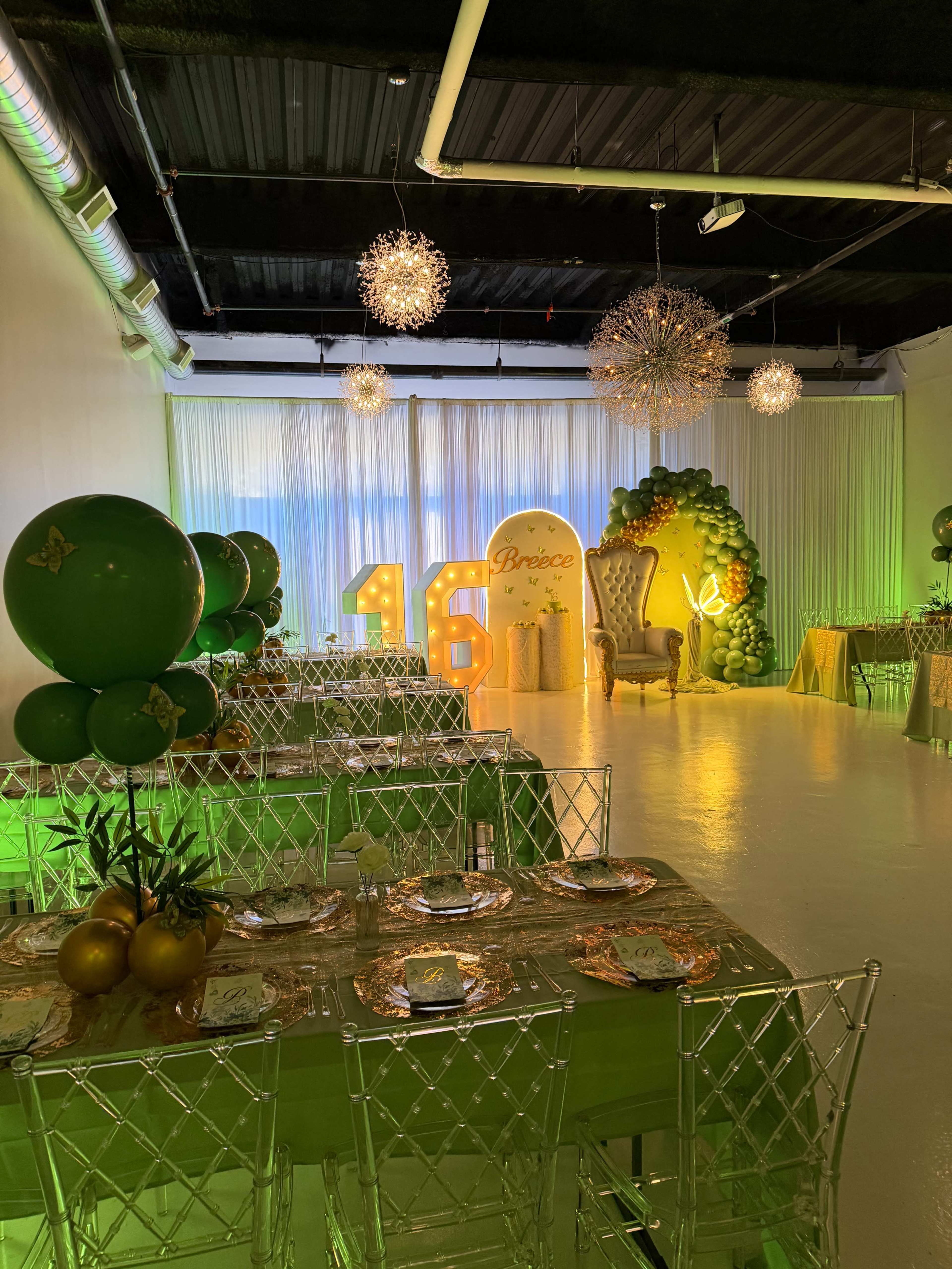The image shows a decorated event space for a 16th birthday celebration, featuring green and gold balloons, elaborate table settings, and a large decorative number "16."