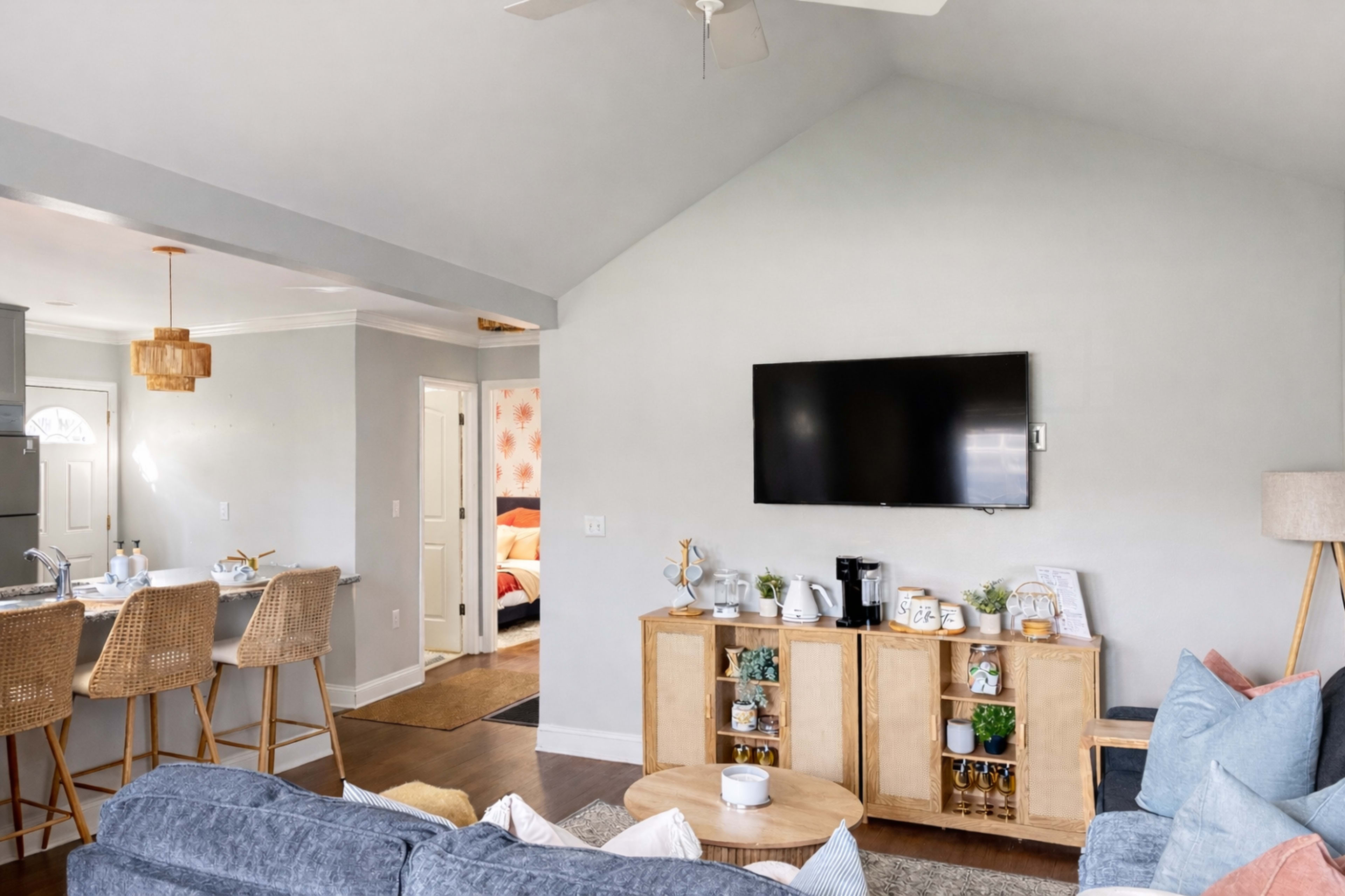 A cozy living room features a blue couch, wooden coffee table, and a wall-mounted television, with a dining area and kitchen visible in the background.