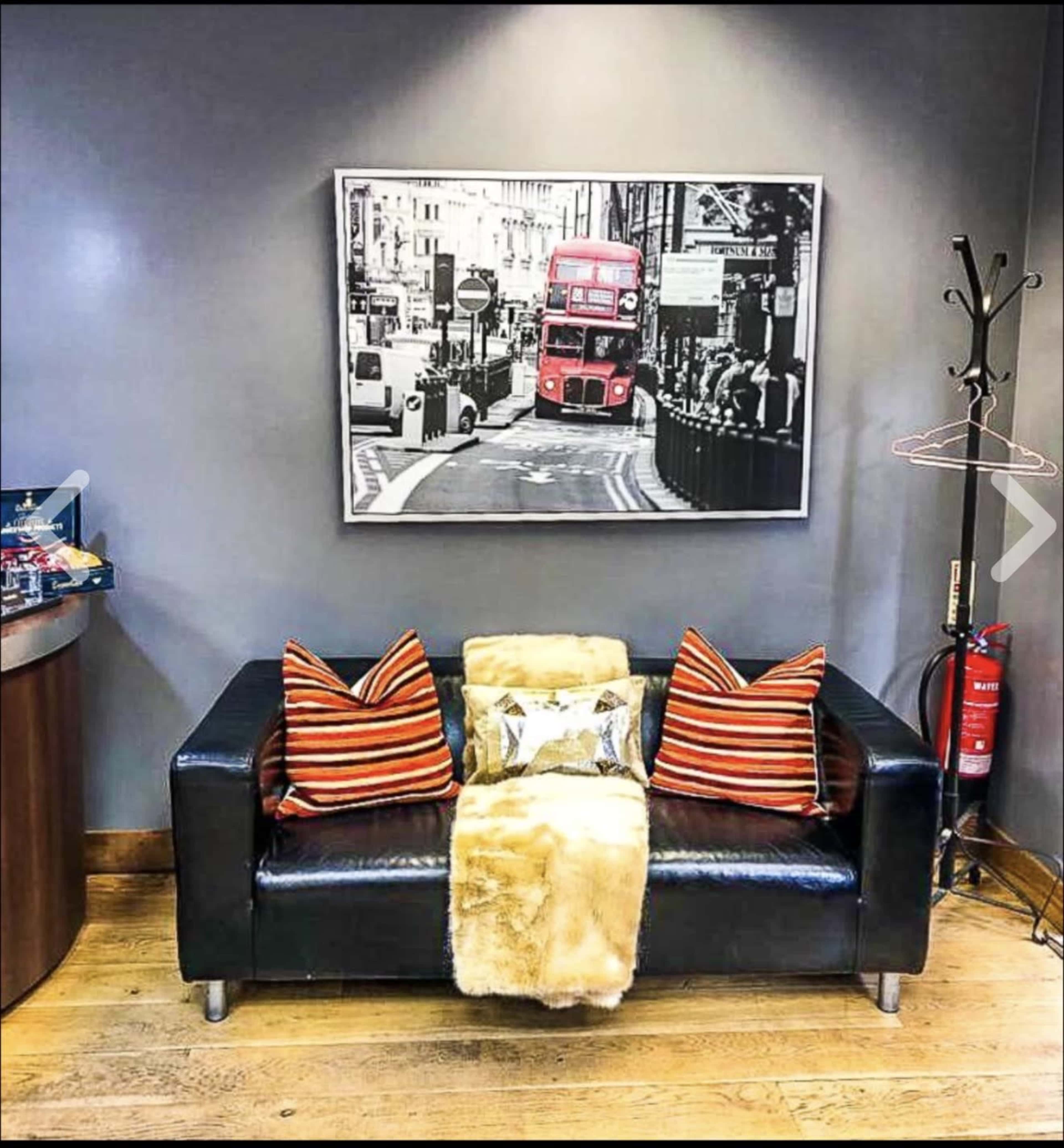 The image shows a black couch adorned with striped pillows and a faux fur throw, positioned against a gray wall featuring a framed black-and-white photograph of a city street with a red double-decker bus.