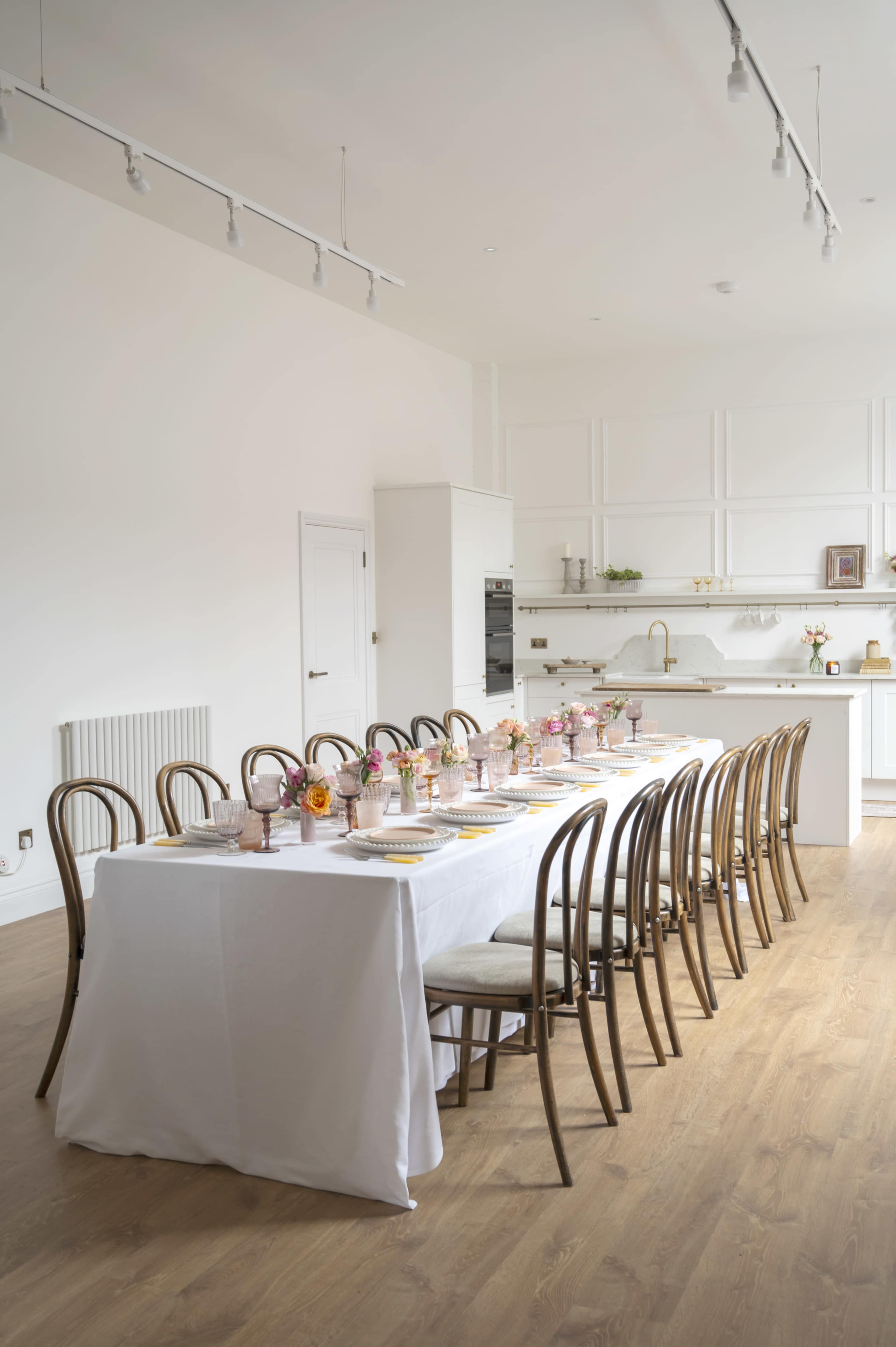 A long dining table is set with flower centerpieces and pink glasses in a bright, modern kitchen.