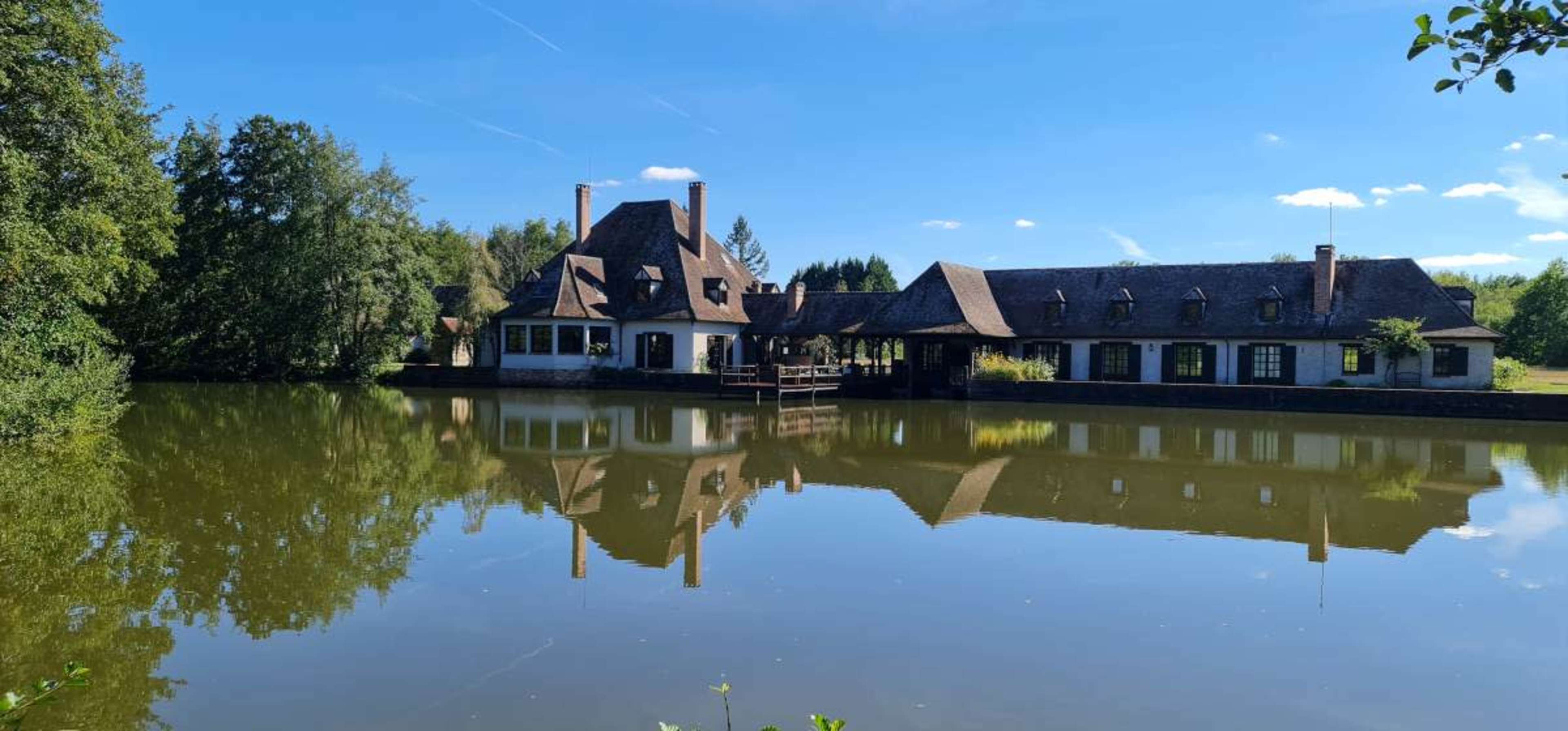The image shows a house with a thatched roof beside a calm pond, surrounded by trees under a clear blue sky.