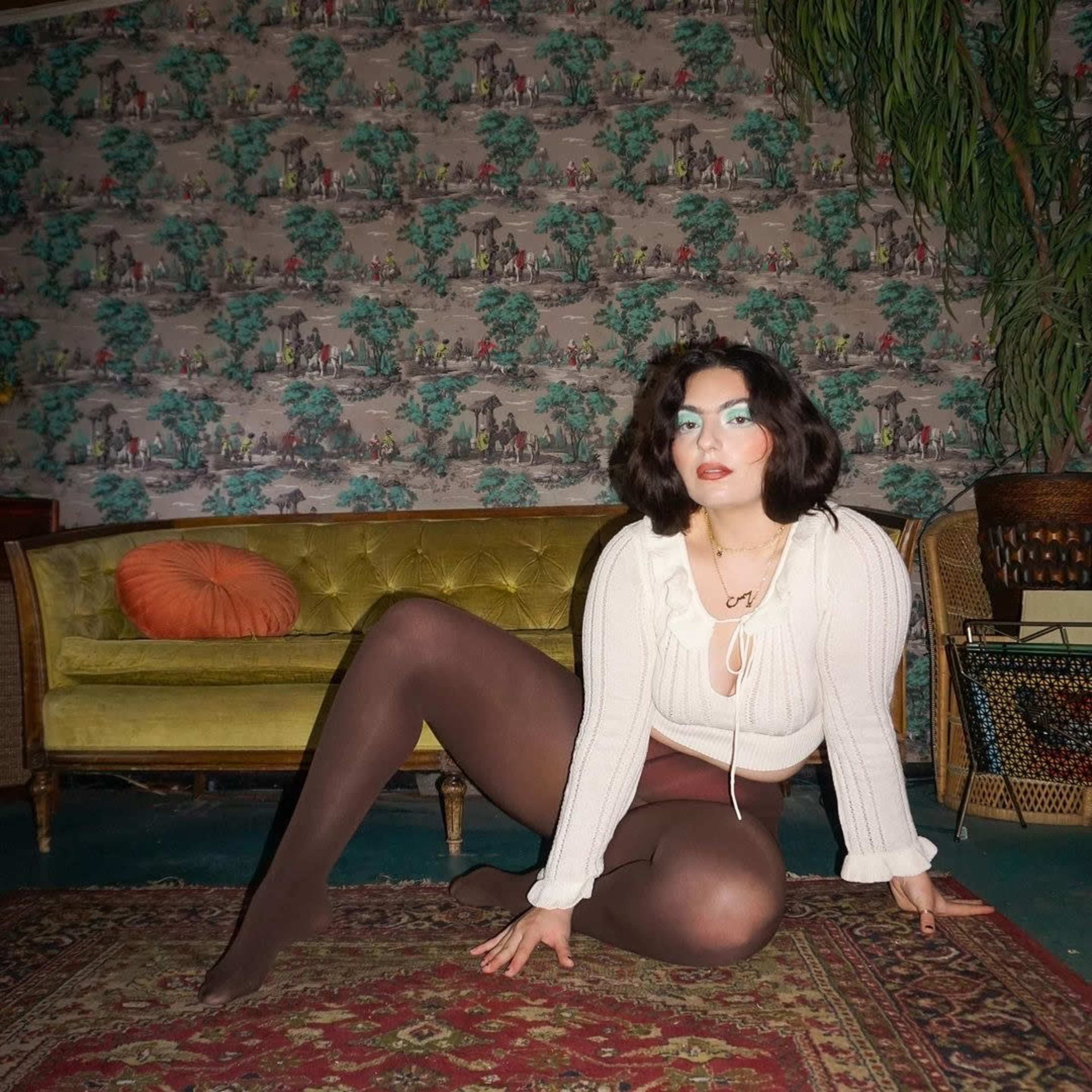 A person with dark hair sits on a patterned rug in front of a vintage couch, wearing a white top and tights, with colorful makeup and a patterned wall behind them.