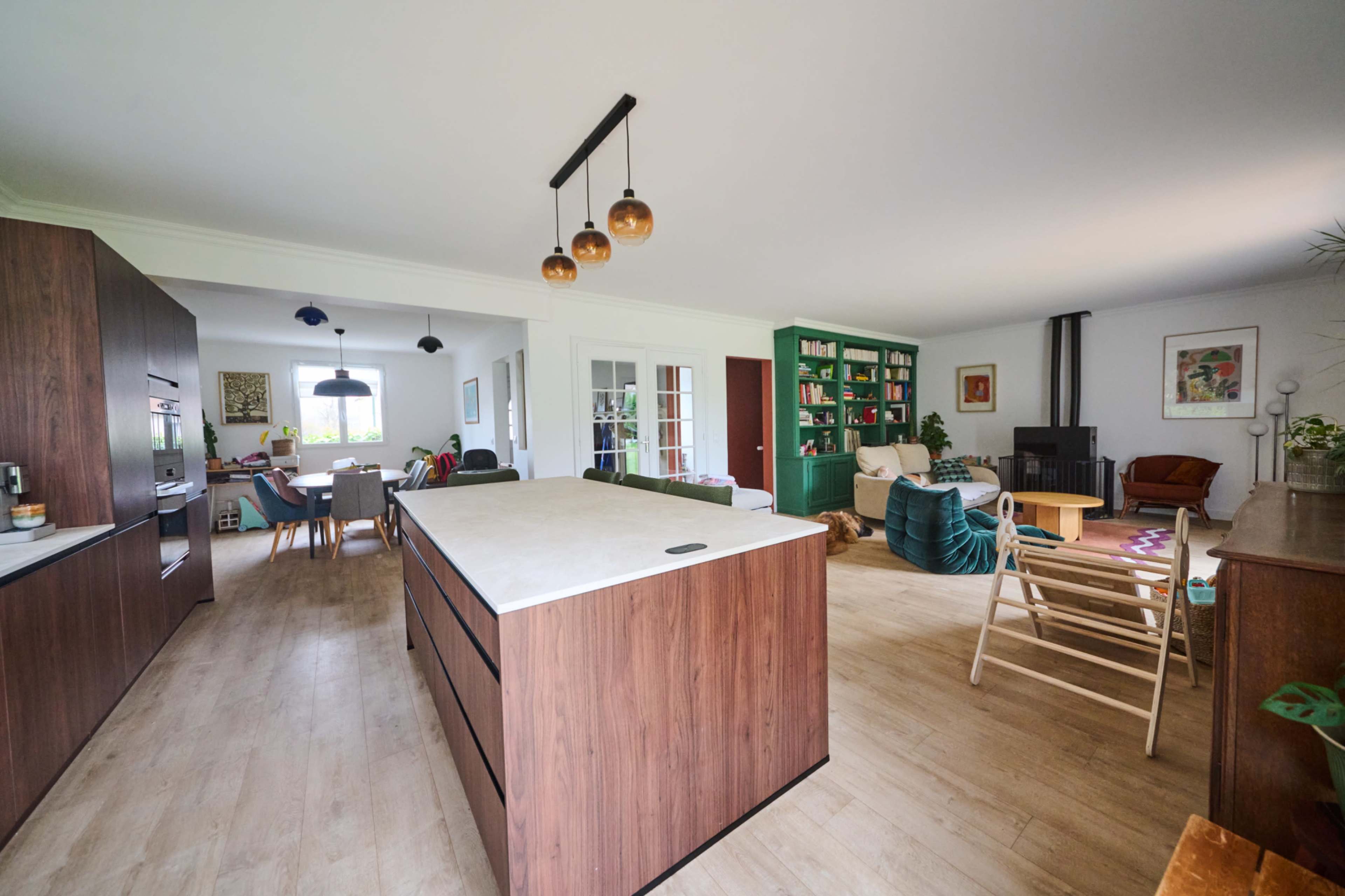 The image shows a modern open-concept living space featuring a kitchen island, dining area, and a cozy sitting area with bookshelves and various seating options.