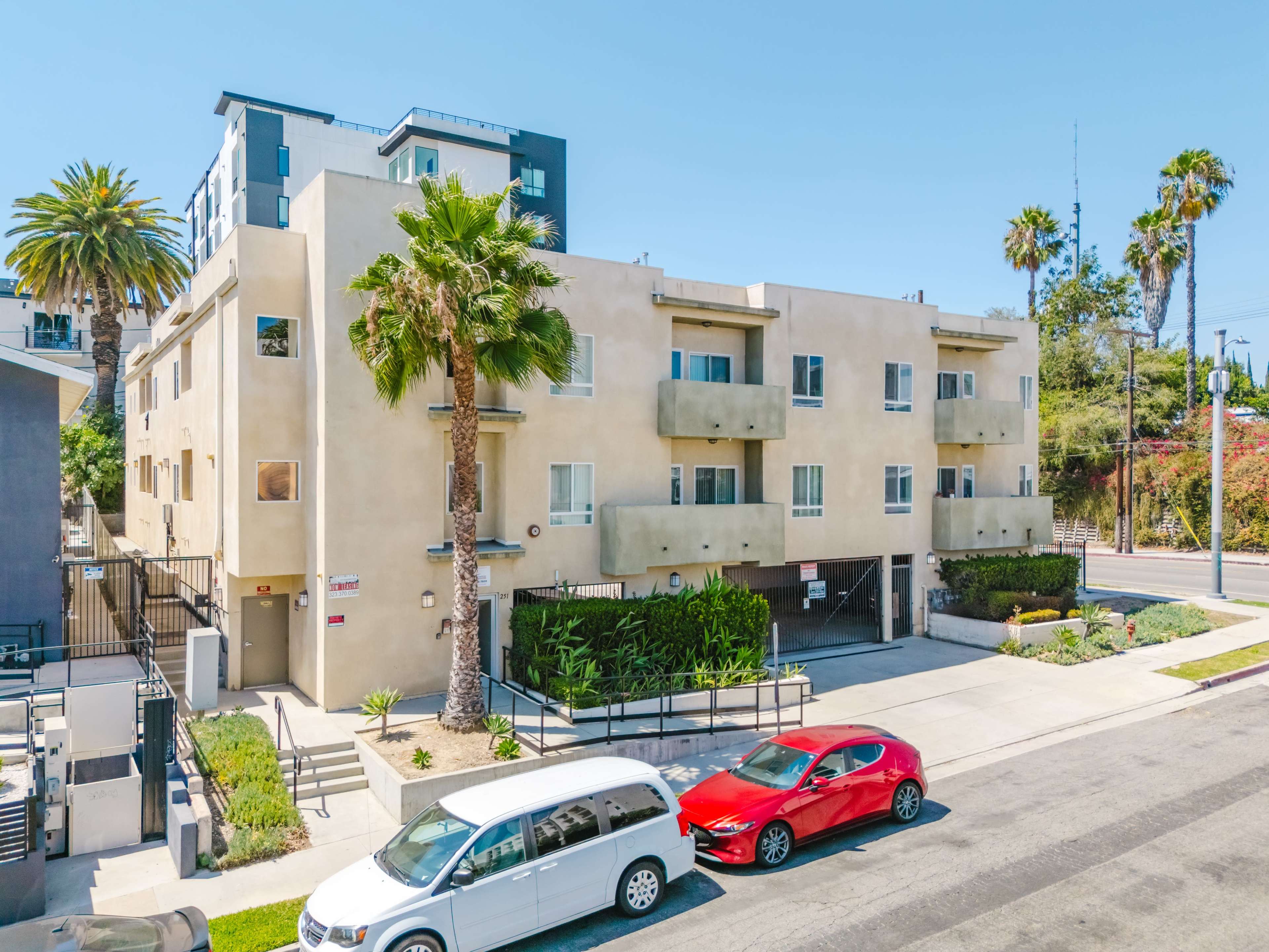A modern, multi-story apartment building with palm trees is seen along a street, featuring parking spaces and landscaped areas.