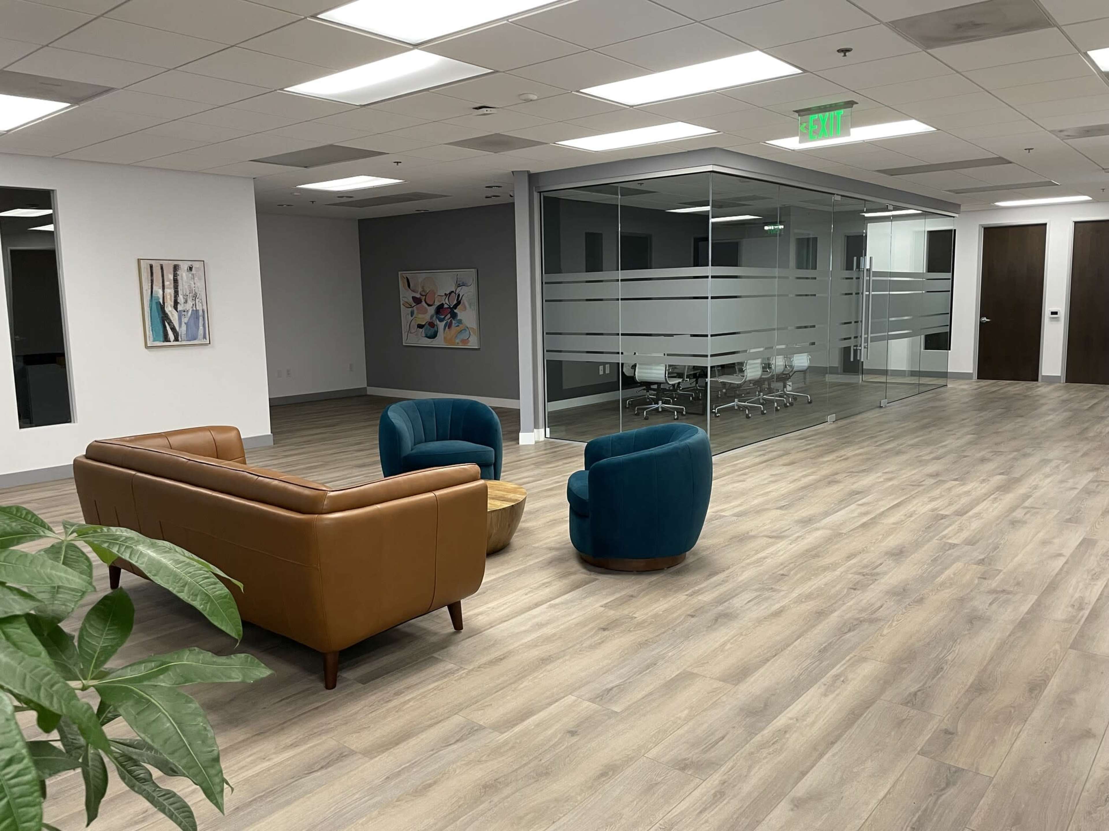 The image shows a modern office space featuring a seating area with a brown leather couch and two colorful chairs, alongside a glass-walled conference room.