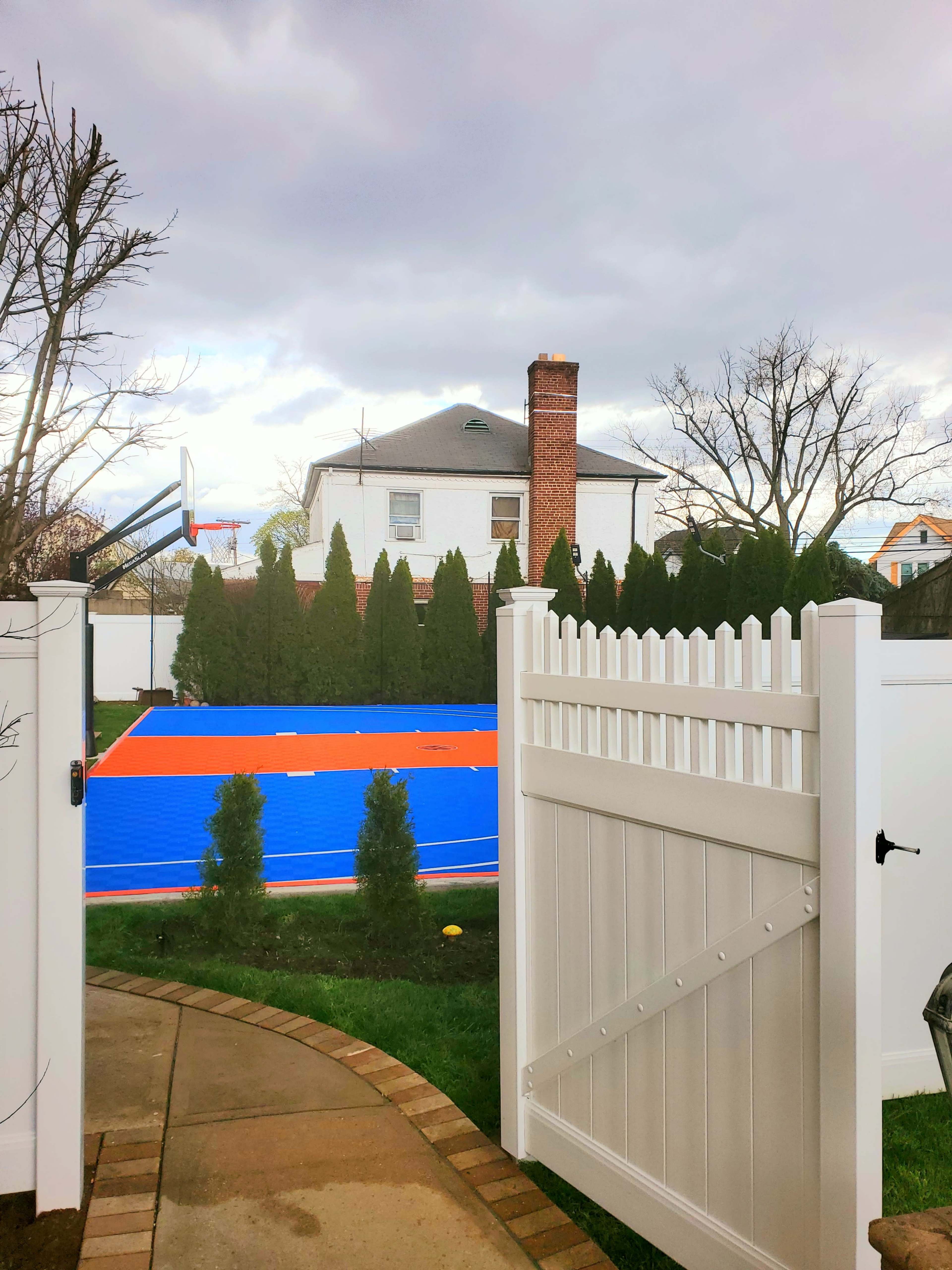 A gated entrance opens to a backyard with a bright blue basketball court and a two-story house in the background.