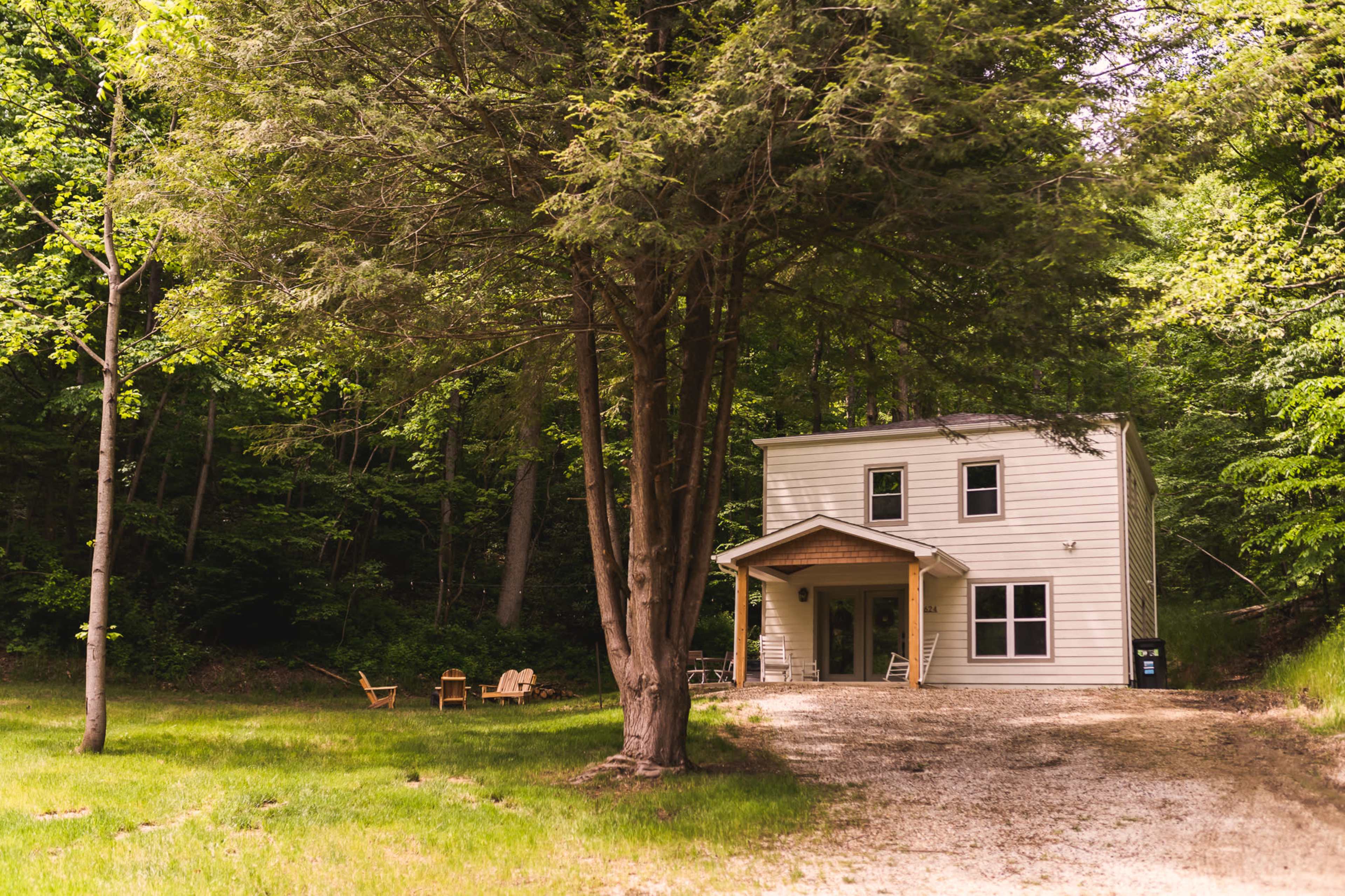 A two-story house with a porch is situated beside a gravel driveway and surrounded by trees in a forested area.