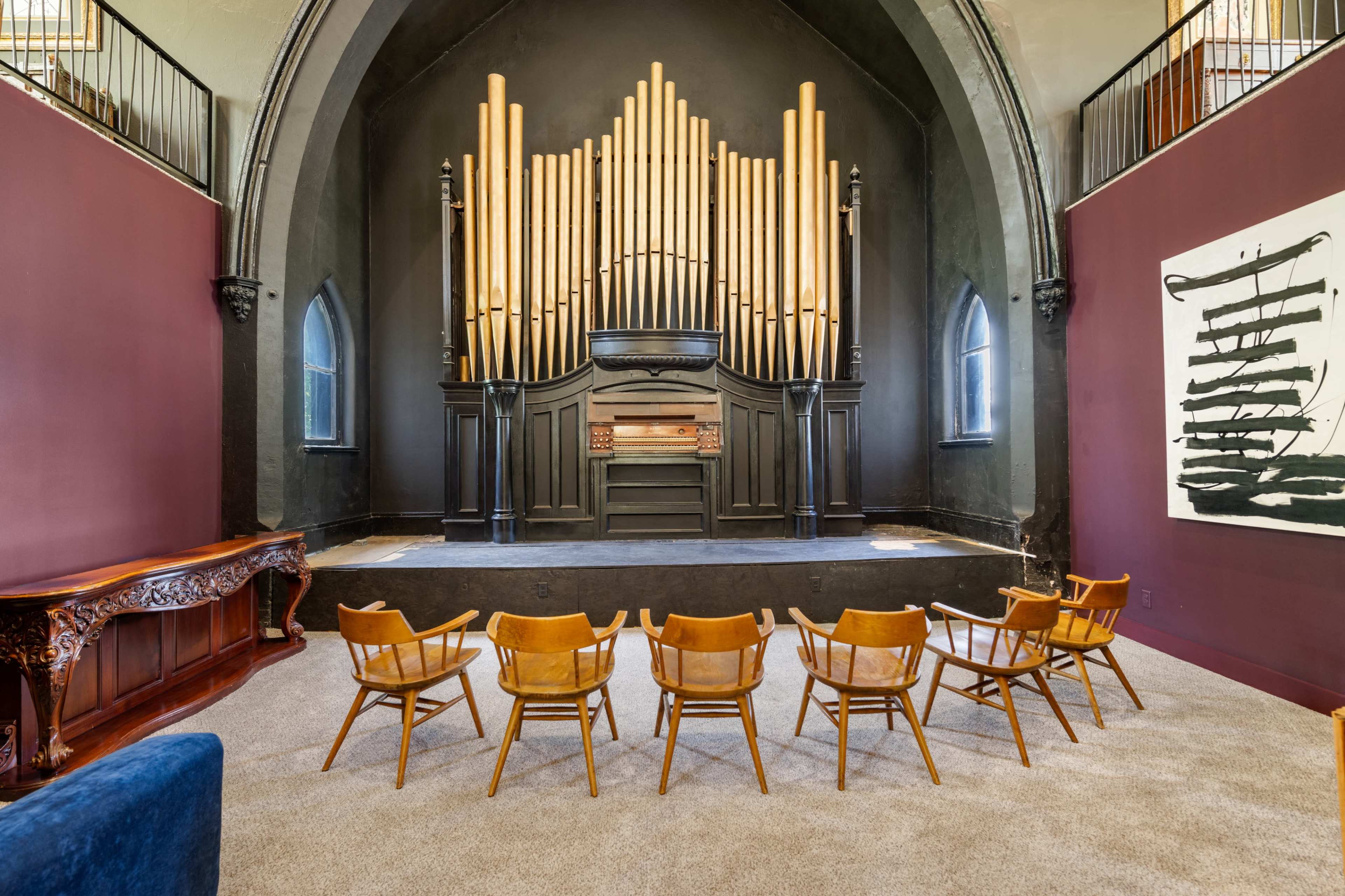 The image shows a performance space featuring a large pipe organ at the back, wooden chairs arranged in a semicircle in front, and dark brown walls with a modern artwork on one side.