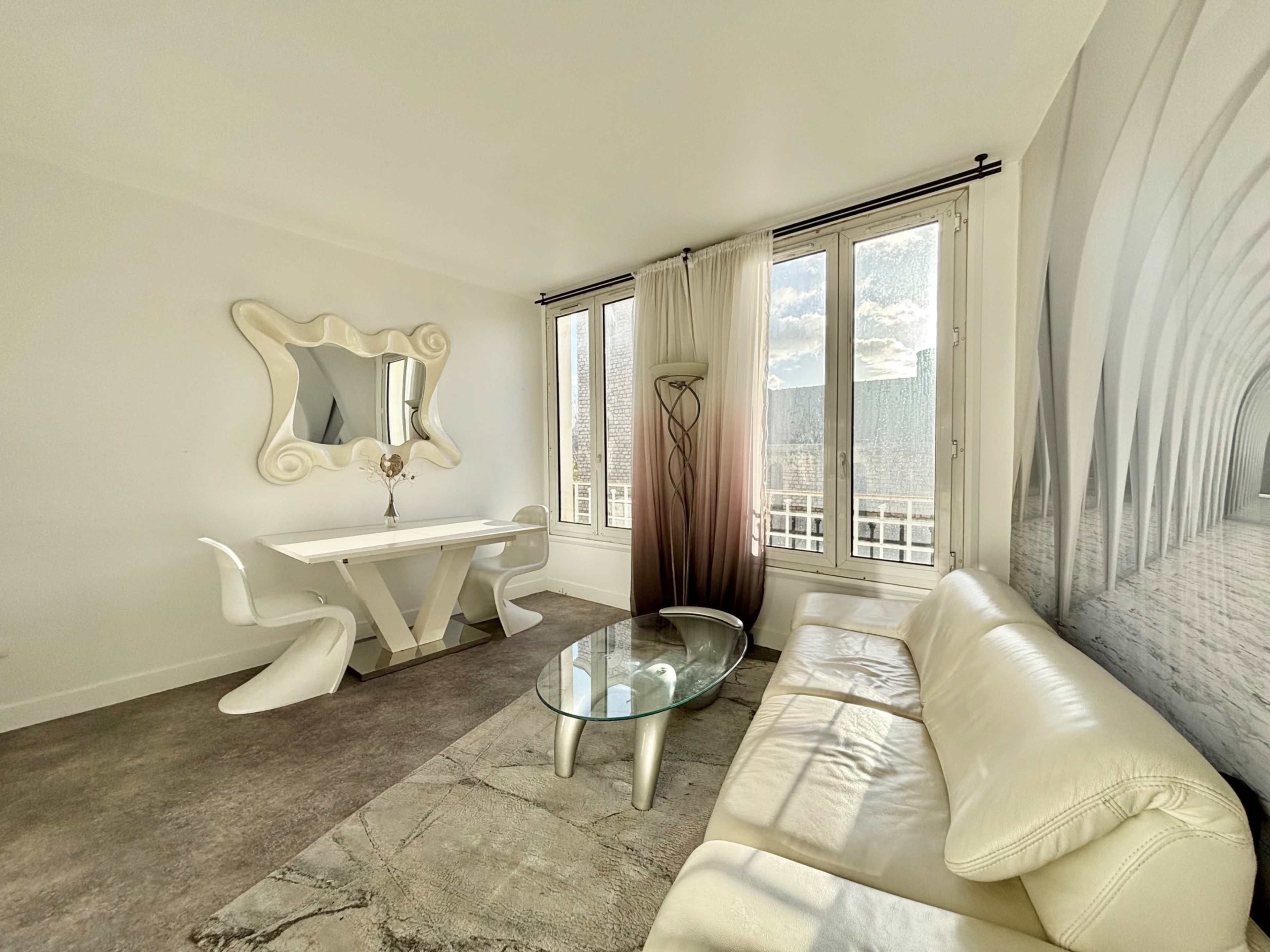 The image shows a modern living room featuring a white leather sofa, a glass coffee table, and a dining area with a unique white table and chairs, illuminated by natural light from large windows.