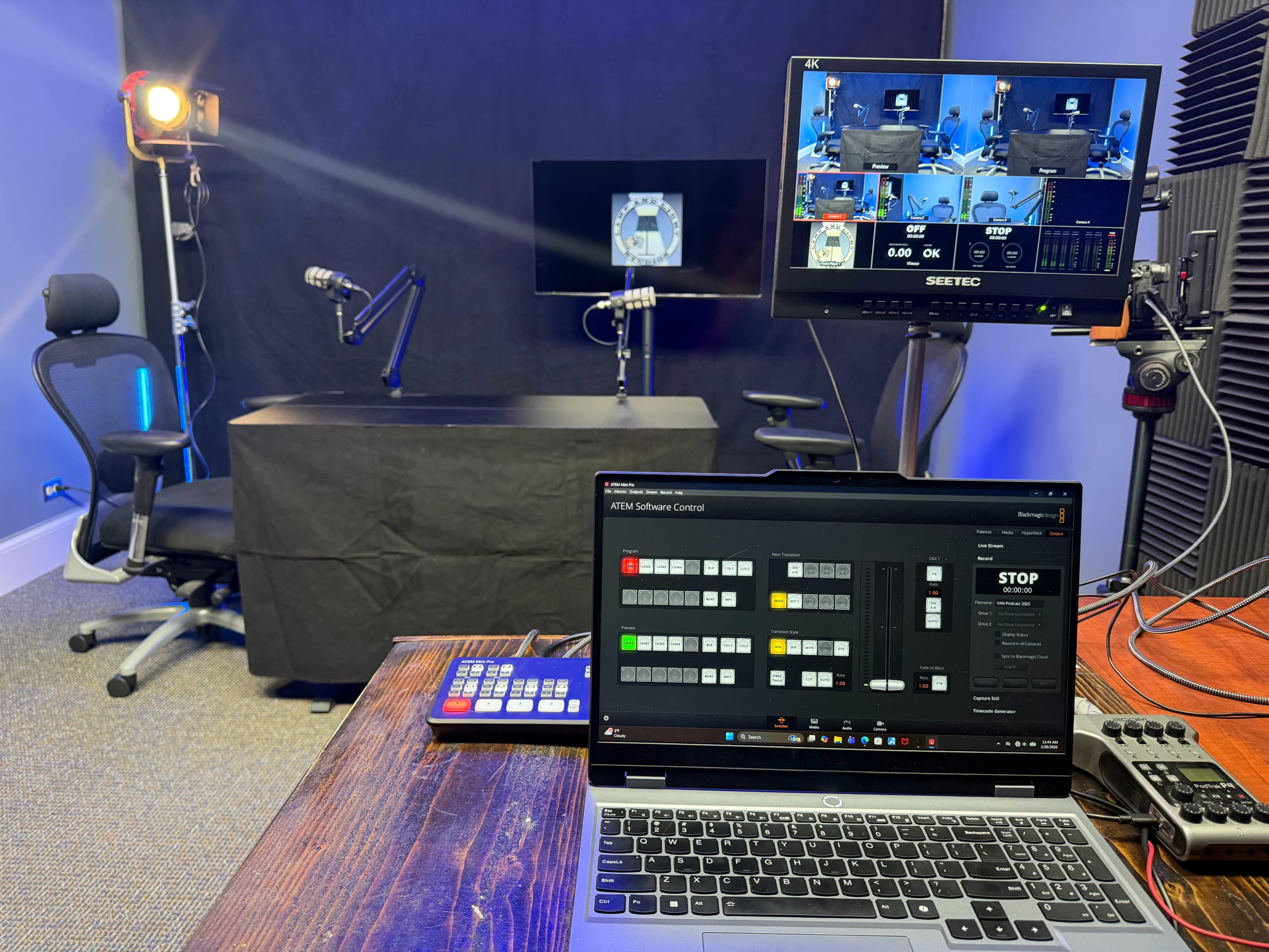 The image shows a video production setup featuring a desk and chair, studio lighting, multiple screens displaying video feeds, and a laptop with control software.