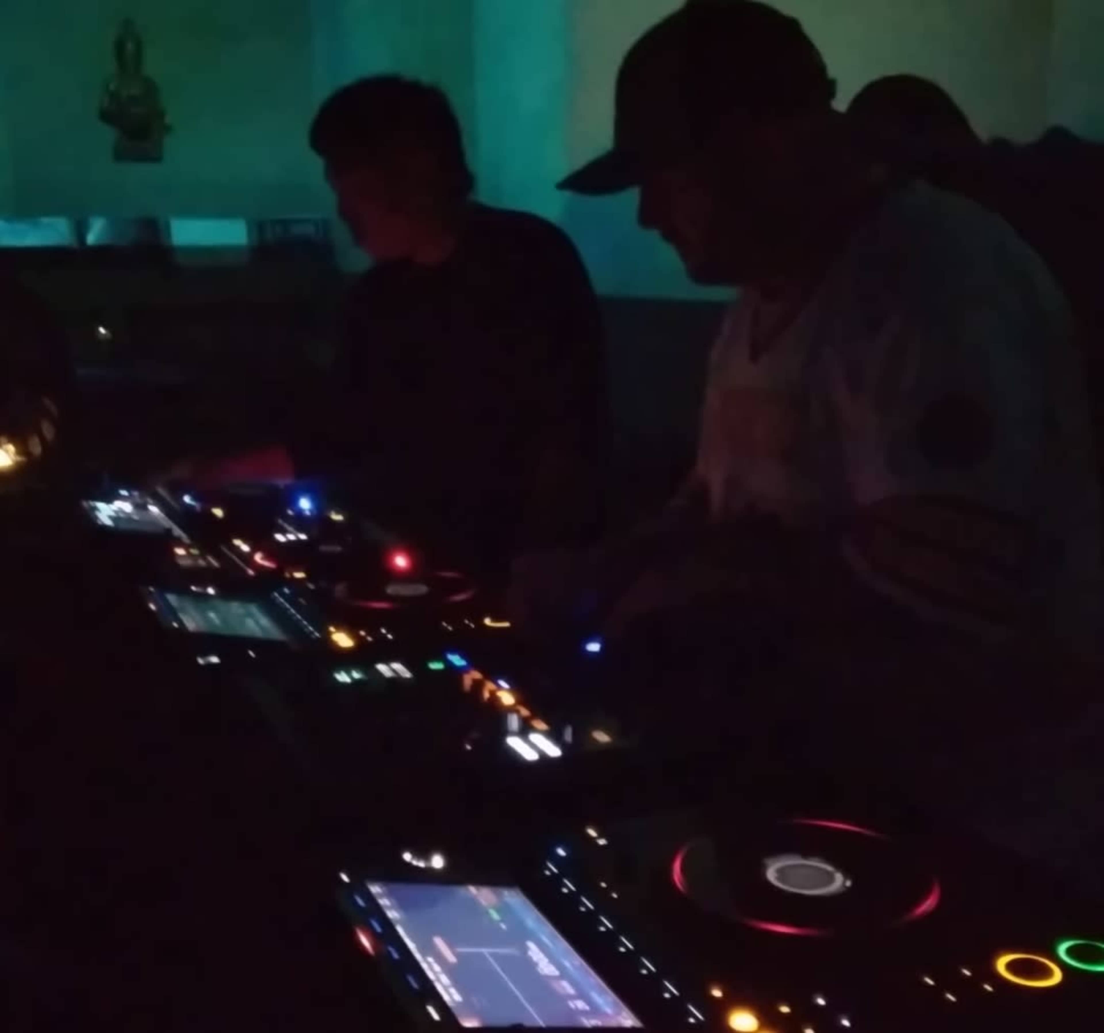 Two DJs are performing at a dimly lit venue, using digital equipment with colorful lights illuminating their consoles.