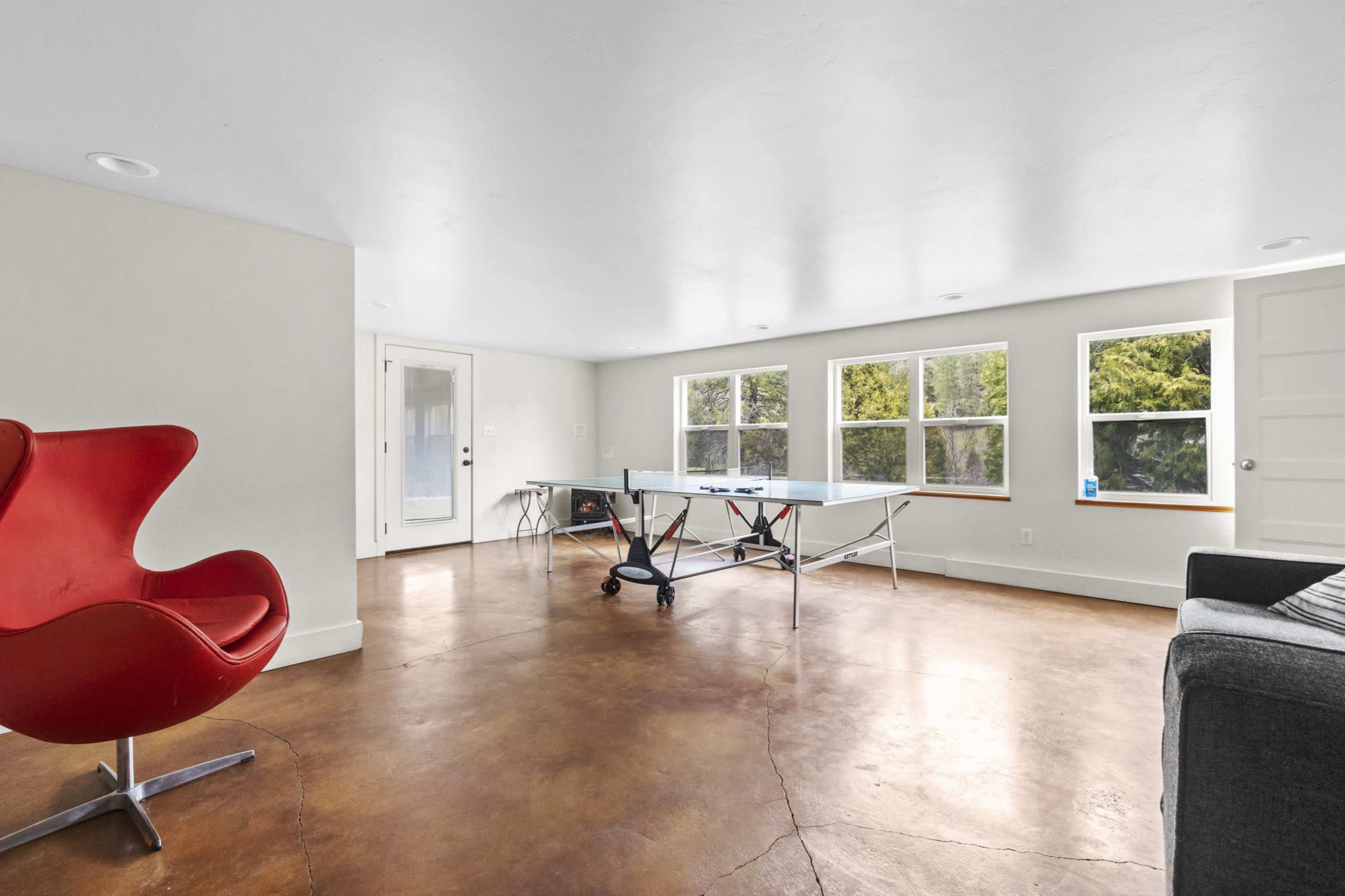 The room features a red chair, a glass table, and large windows allowing natural light, with a concrete floor and white walls.