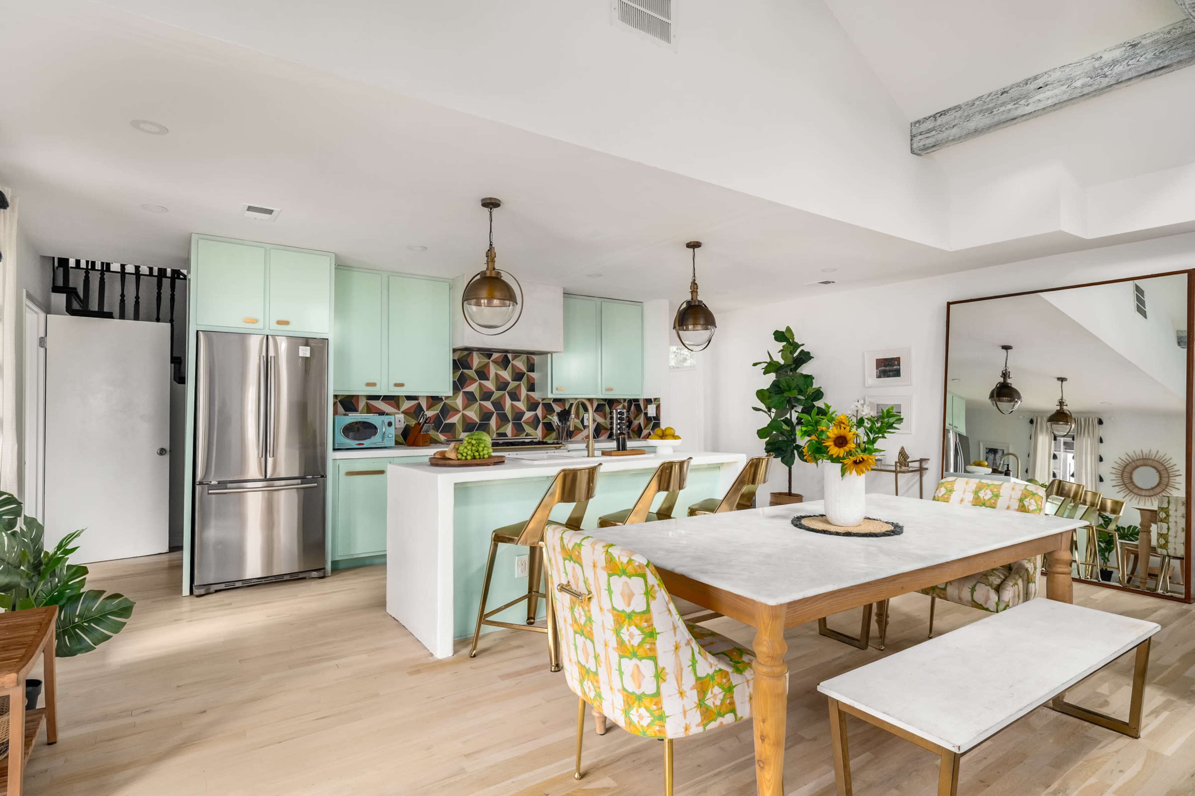 The image shows a modern kitchen and dining area featuring mint green cabinets, a large table with floral-patterned chairs, and bright sunlight streaming through a window.