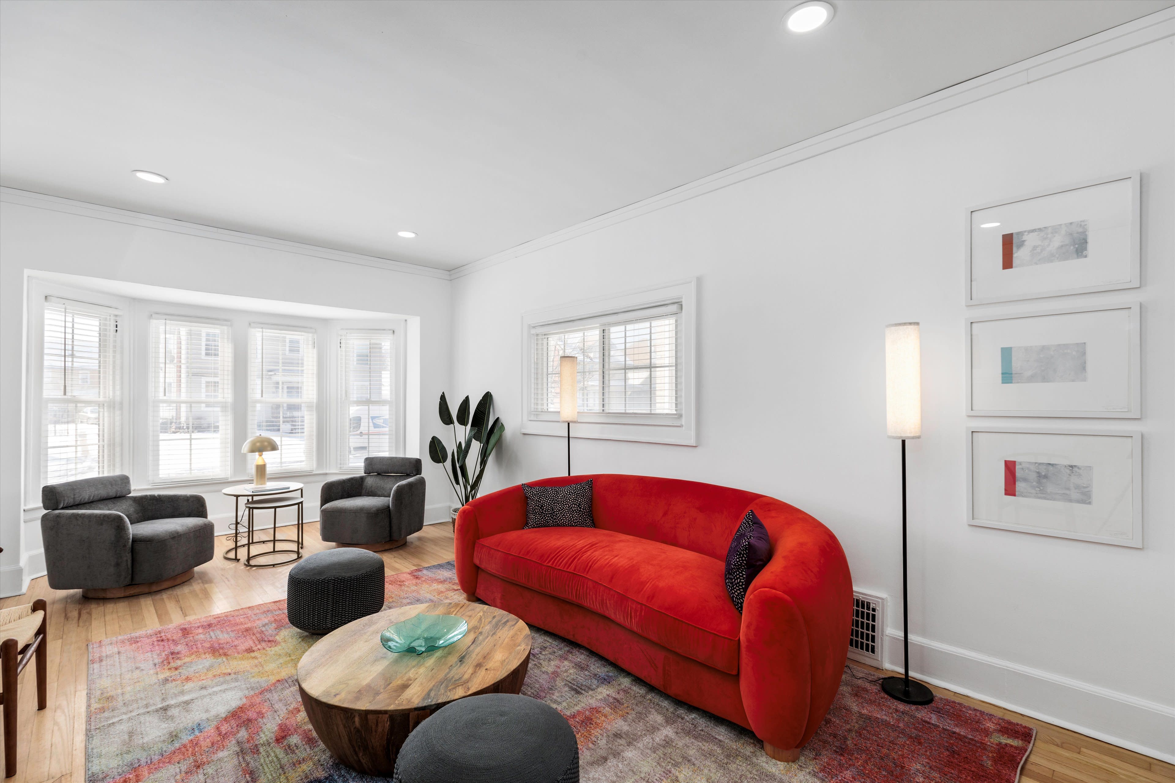 The living room features a red sofa, two gray armchairs, a round wooden coffee table, and natural light flowing through bay windows.