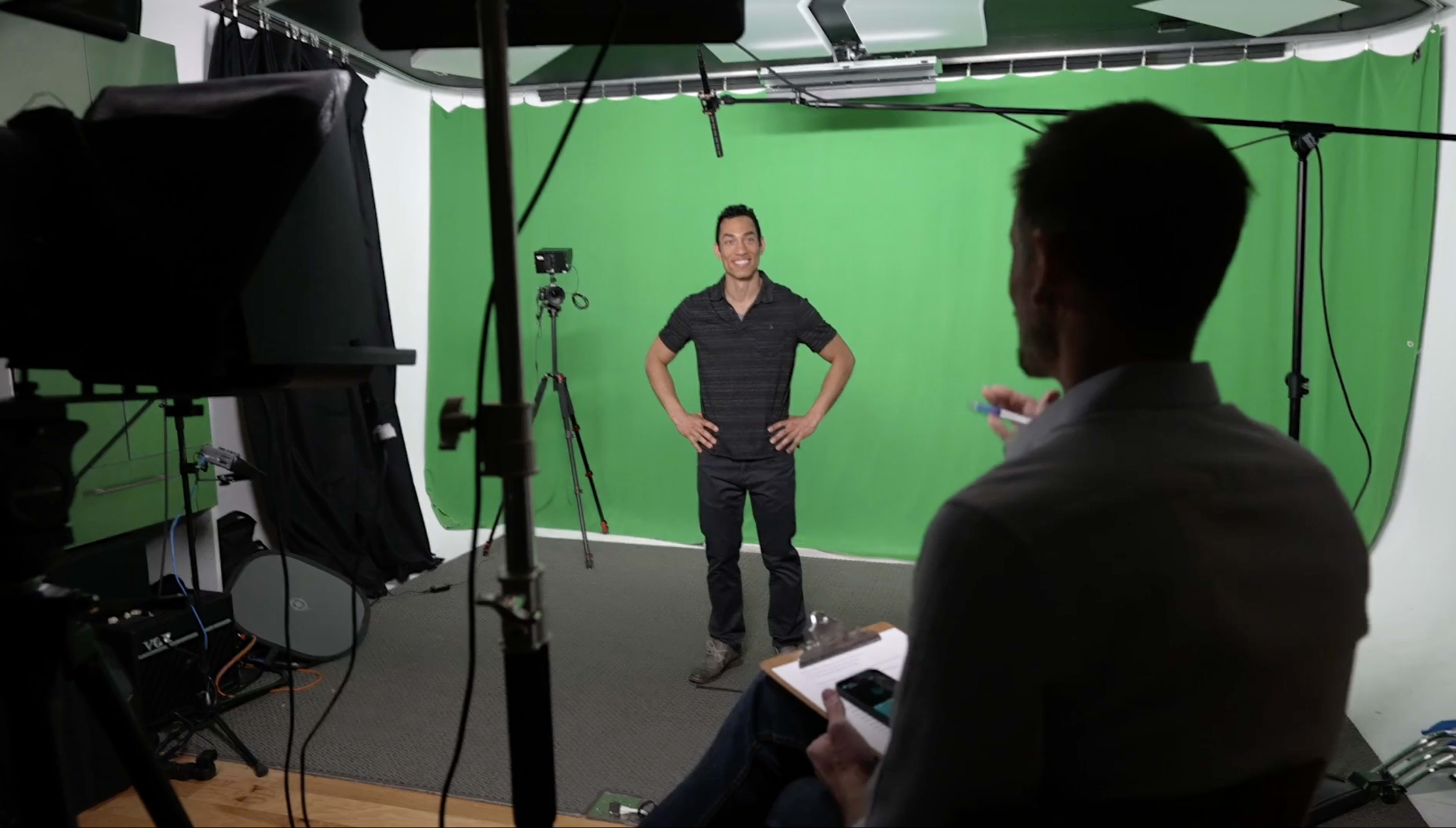 A man stands confidently in front of a green screen while another person sits behind a camera, taking notes.