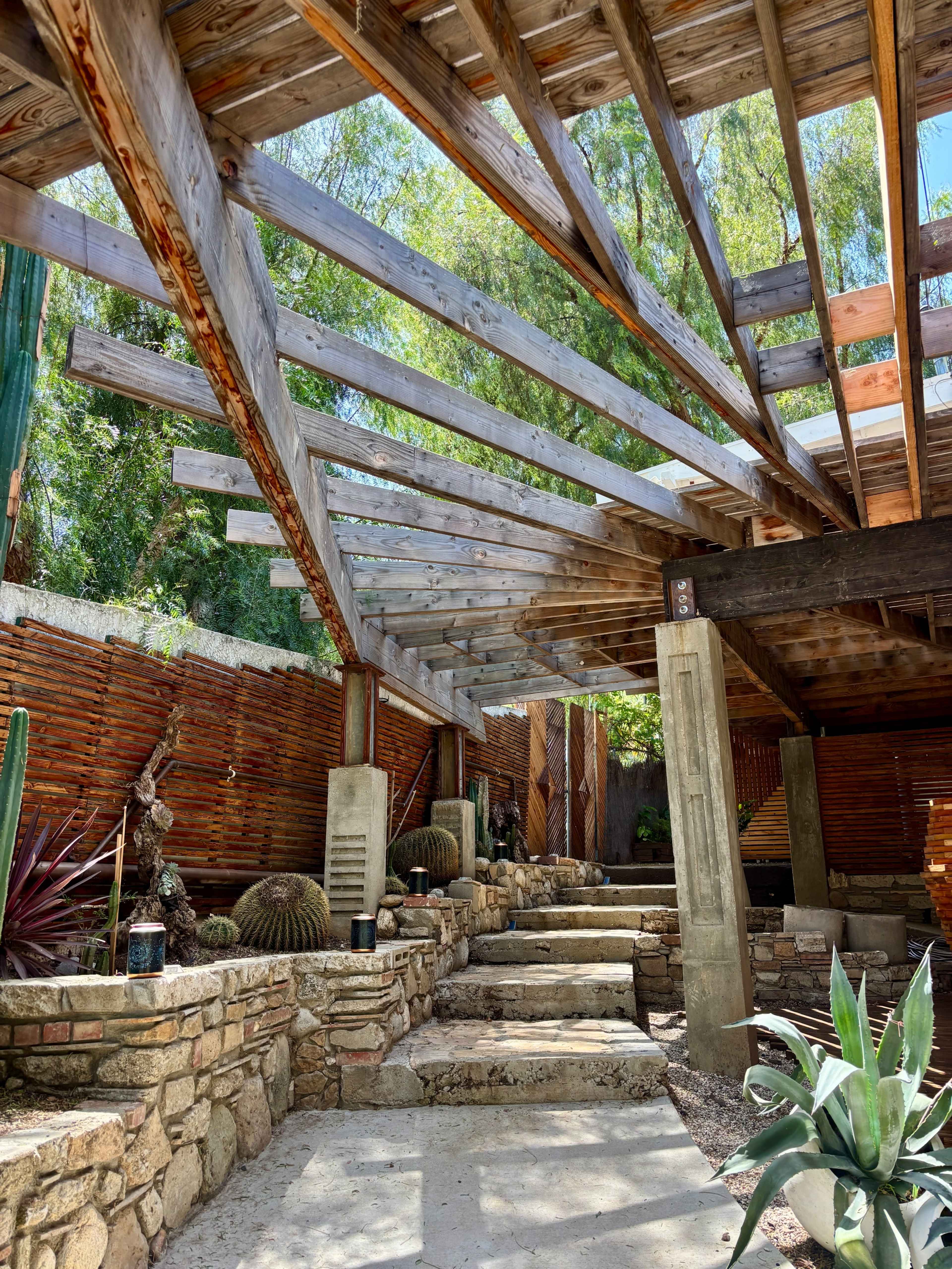 The image depicts an outdoor architectural space with wooden beams, stone steps, and various plants, including cacti and succulents, surrounded by a rustic stone wall.