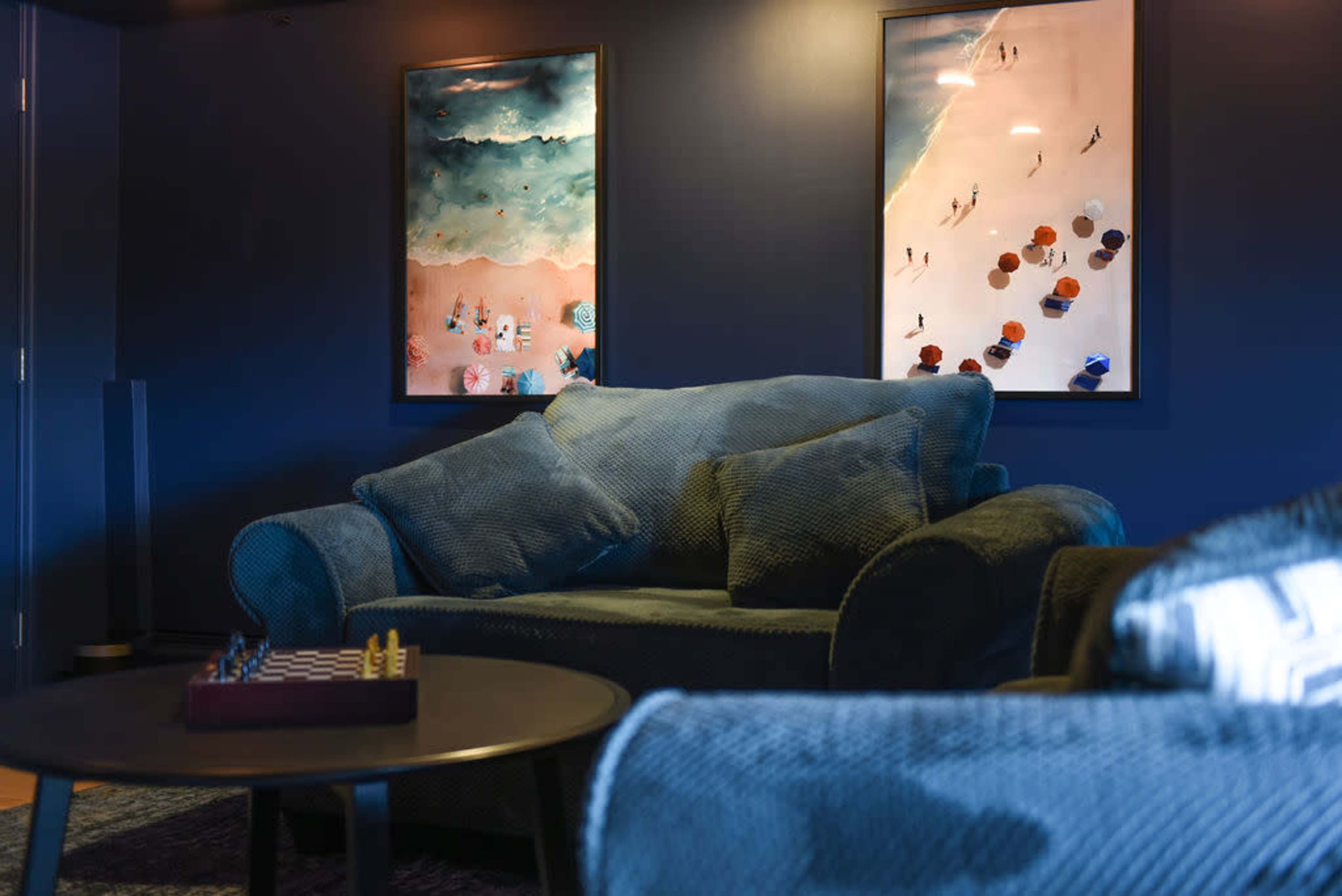 A cozy living room features a blue couch, a round table with a chess set, and two framed beach-themed artworks on the wall.