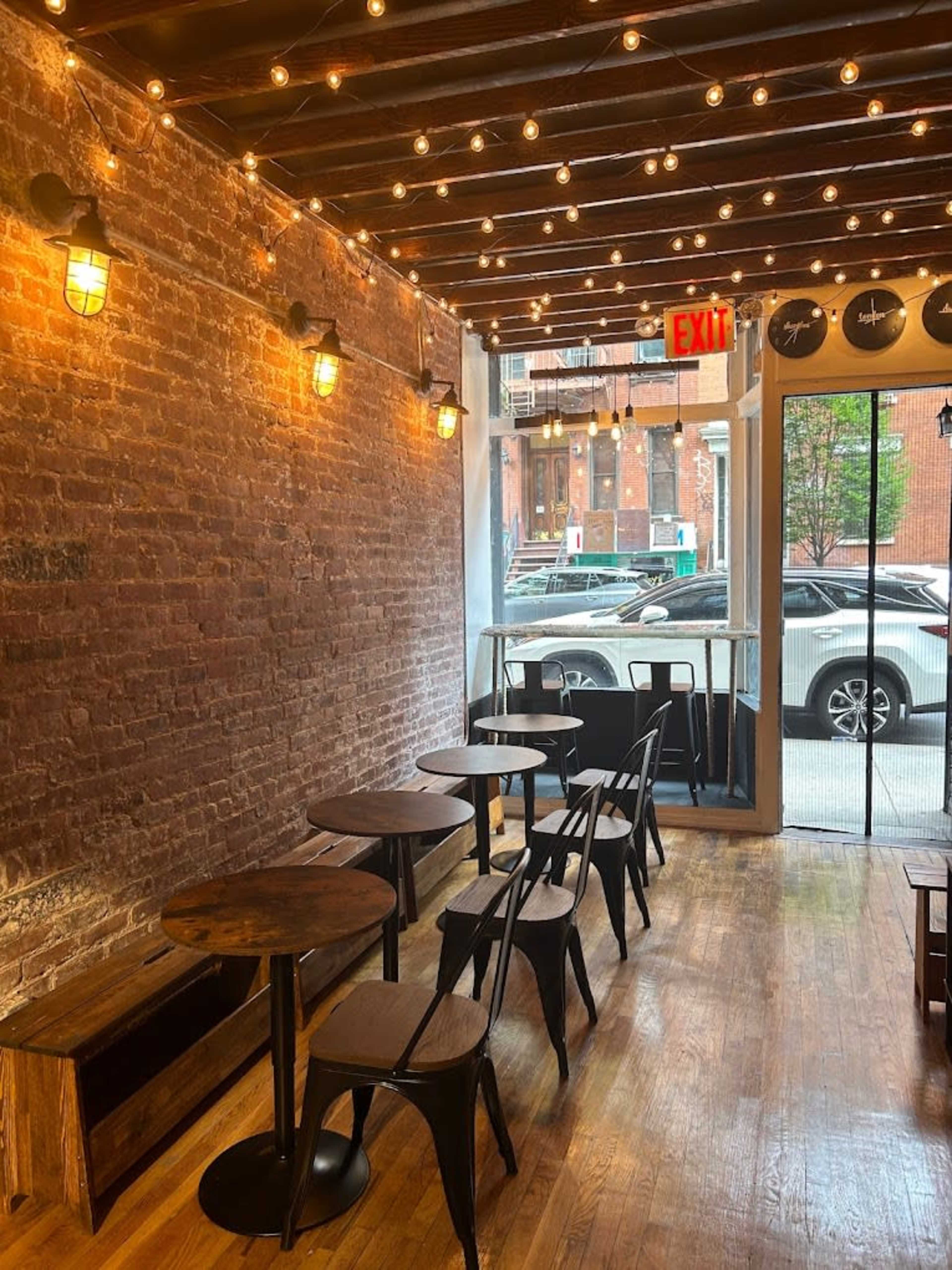 An empty café with wooden tables and chairs, exposed brick walls, and string lights overhead, near a large window displaying a street view.
