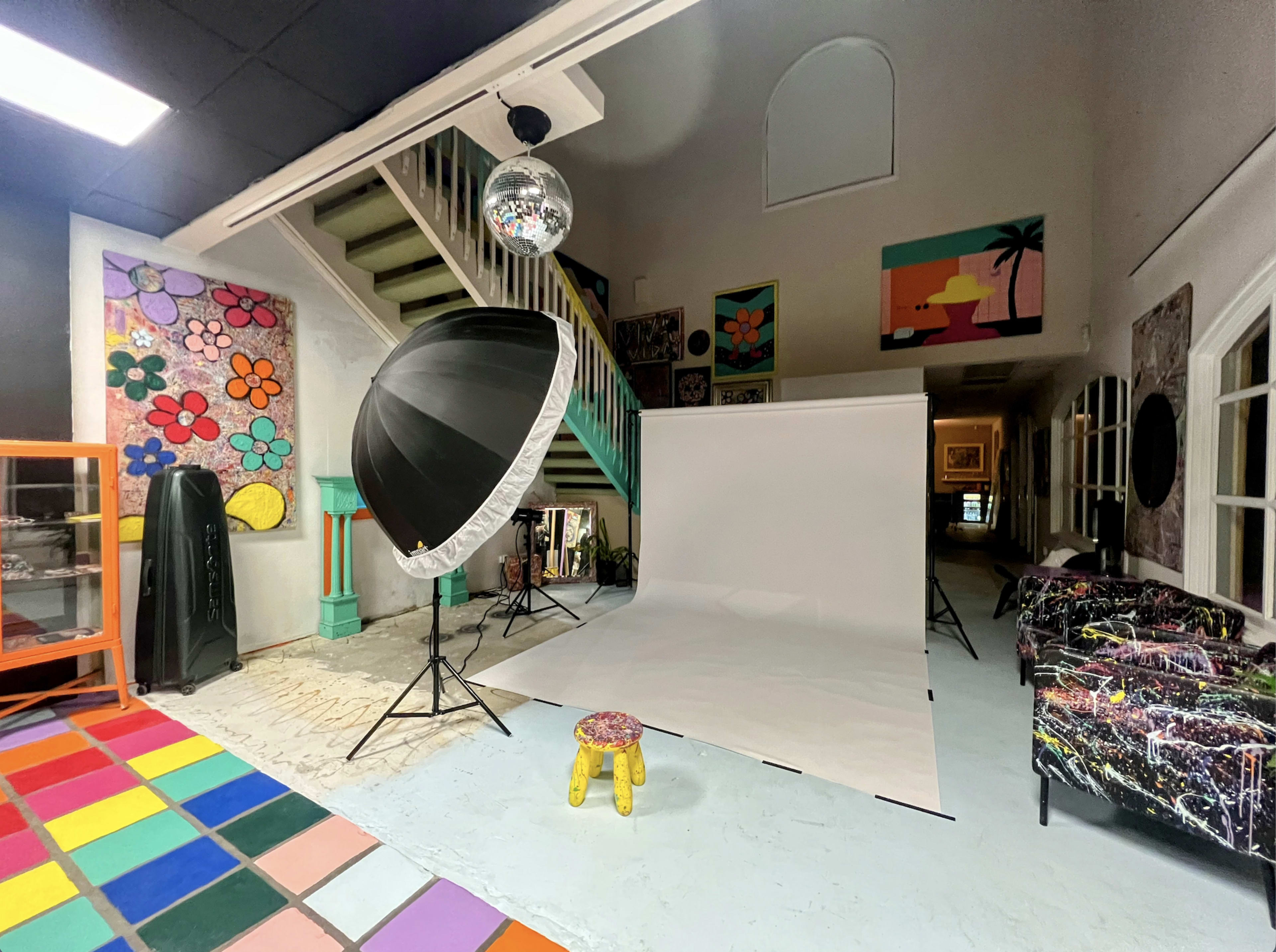 The image shows a colorful studio space with a white backdrop set up for photography, featuring a large umbrella light, a disco ball, and vibrant artworks on the walls.
