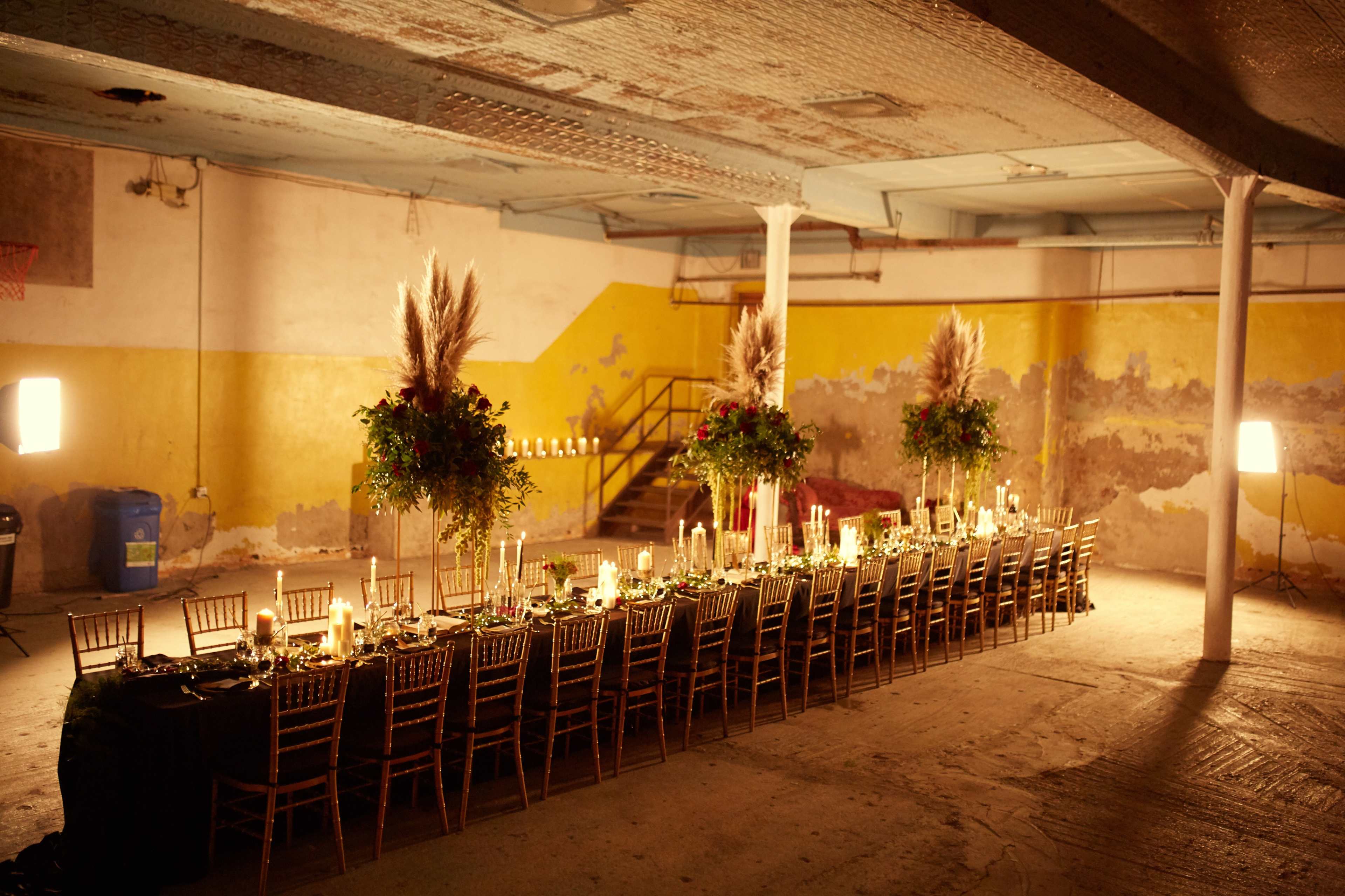 A long dining table is set in an industrial space with yellow walls and dim lighting, adorned with candles and floral arrangements.