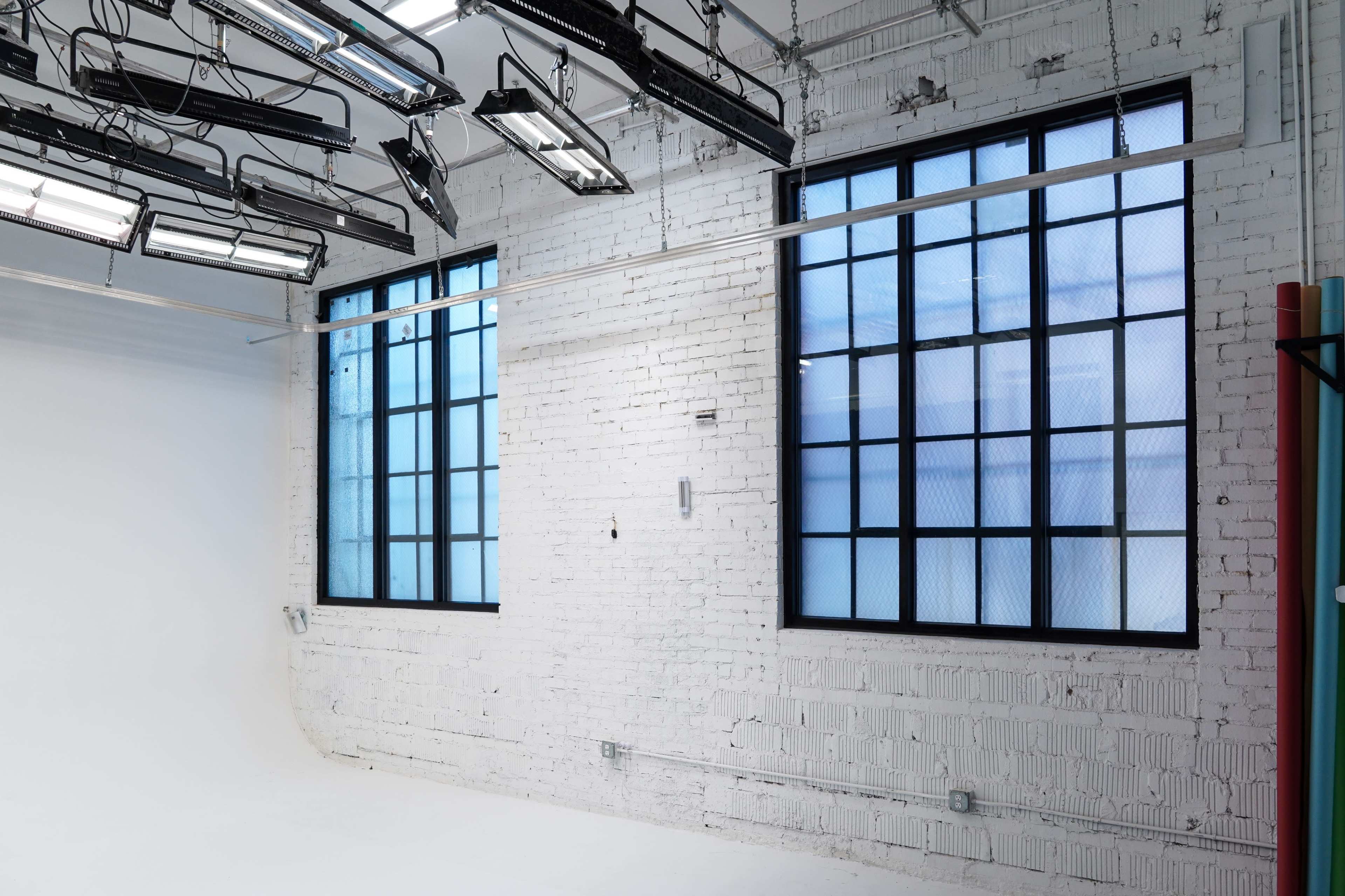 The image shows a bright, minimalist studio space with large windows and exposed brick walls.