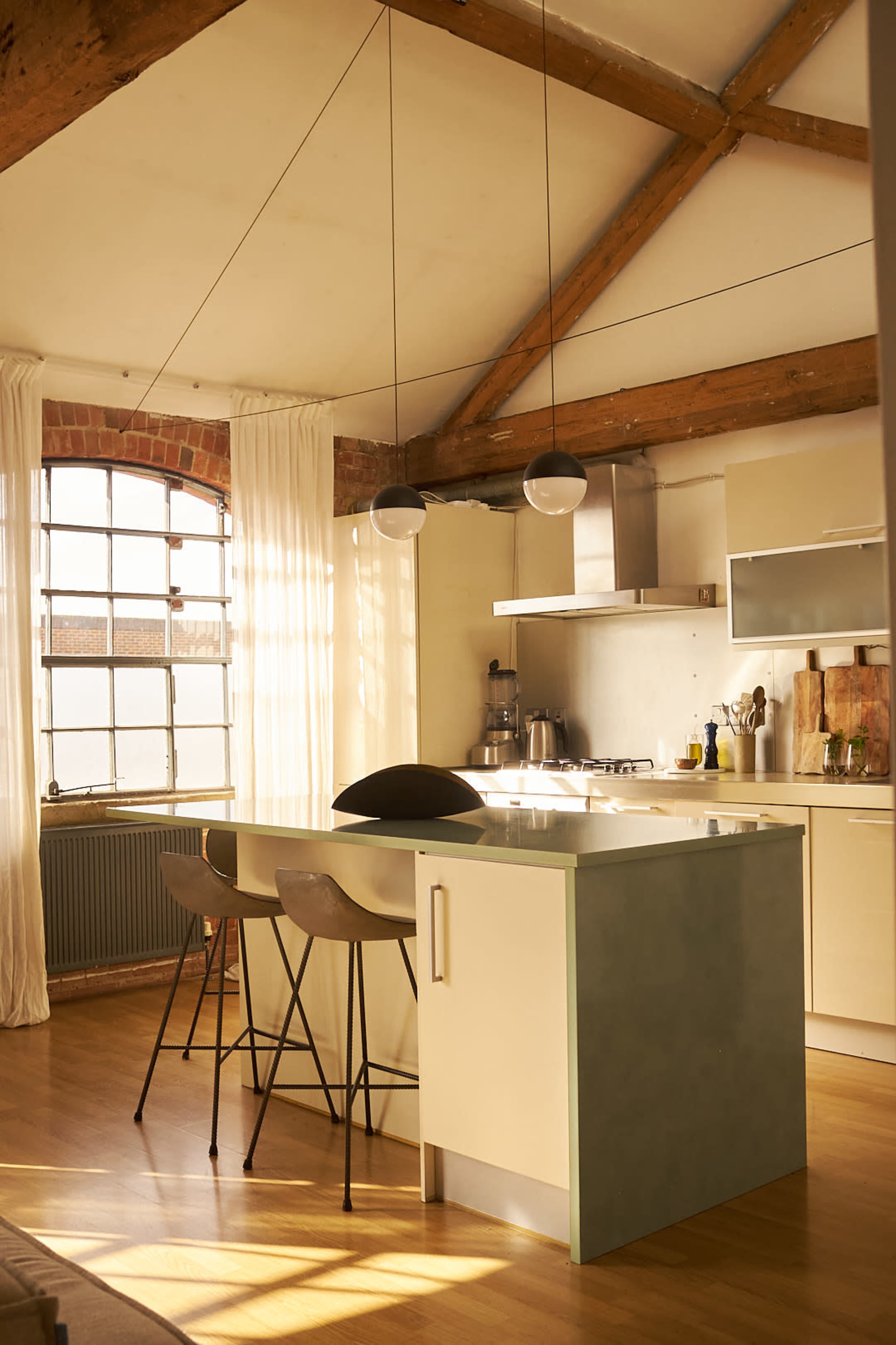 The image shows a modern kitchen with an island and bar stools, featuring large windows and exposed wooden beams.