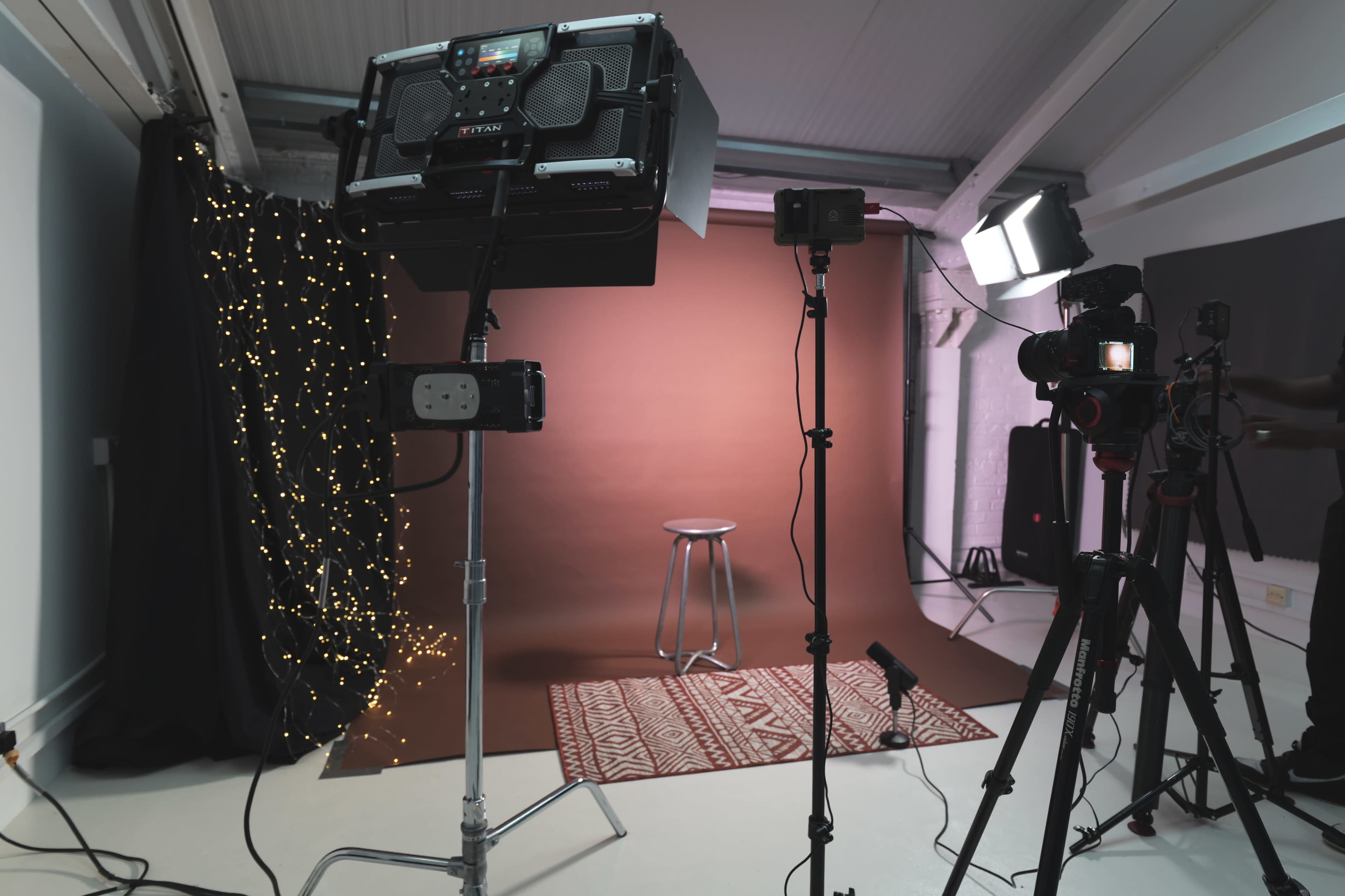 The image shows a photography studio setup with a brown backdrop, a stool, decorative lights, and various cameras and lighting equipment arranged around the scene.