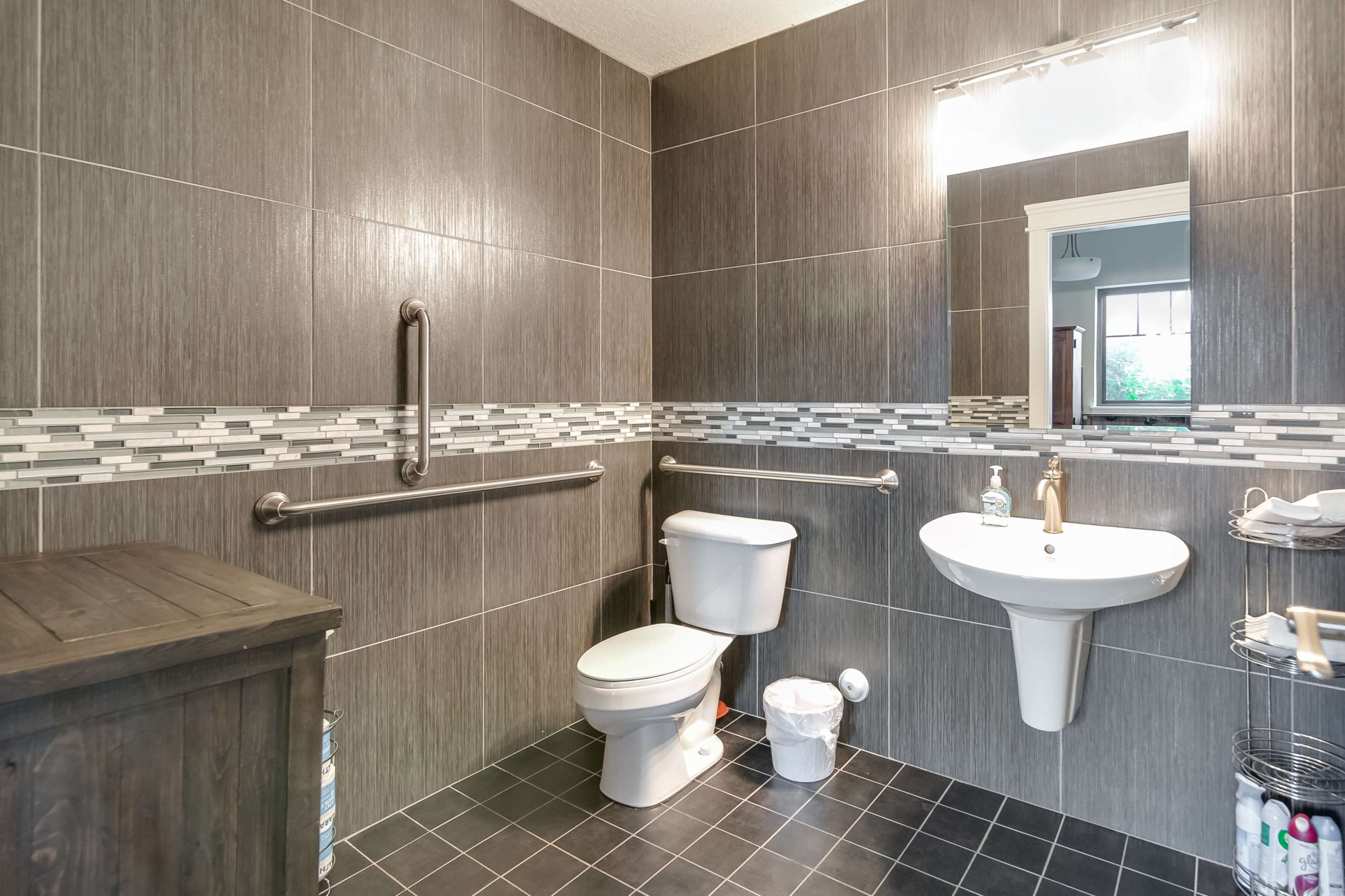 A modern bathroom with gray tile walls, a toilet, a basin with a faucet, and a mirror above it, along with grab bars and a wooden storage unit.