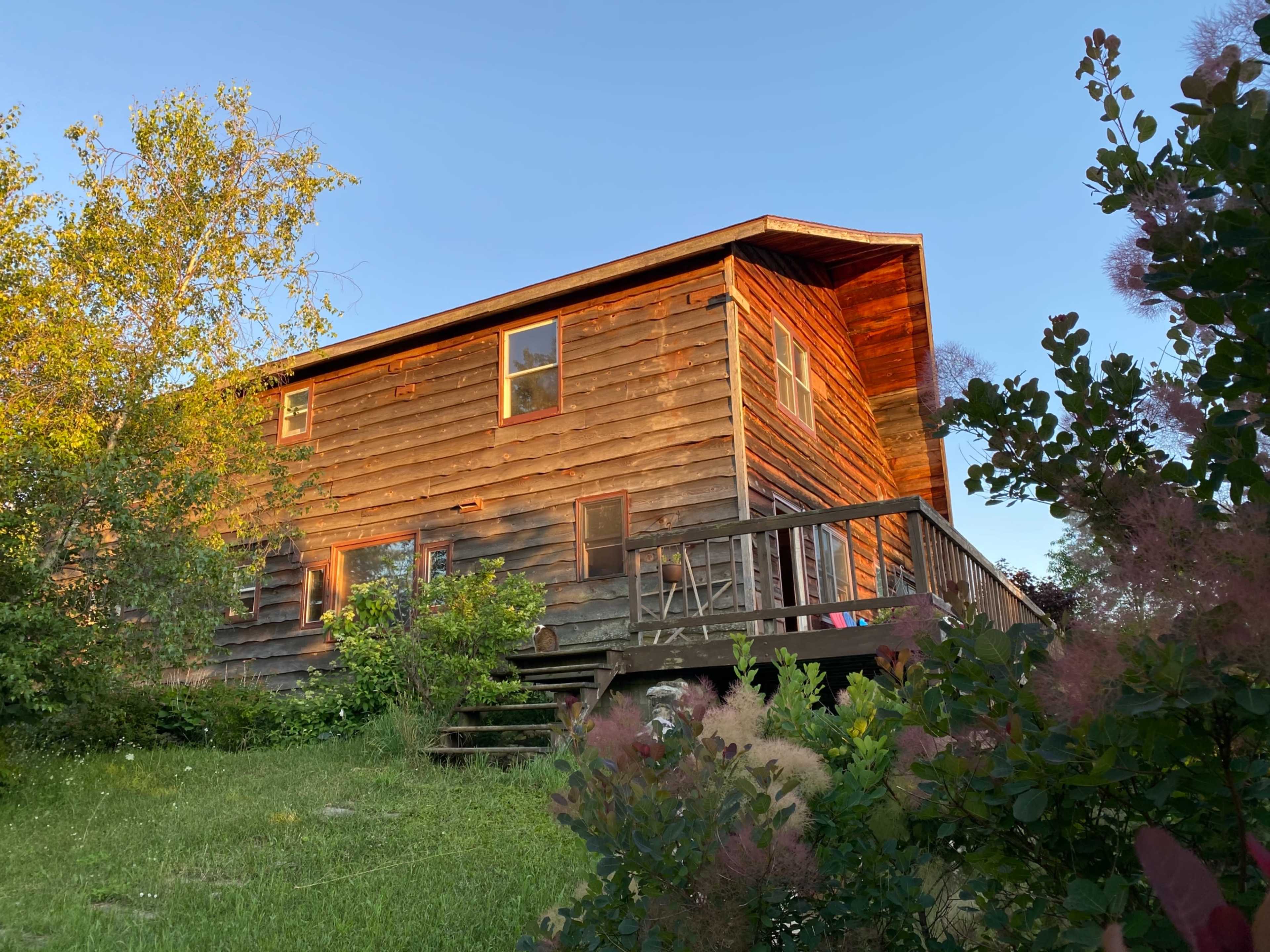A two-story wooden house with a porch is surrounded by greenery and sunlight.