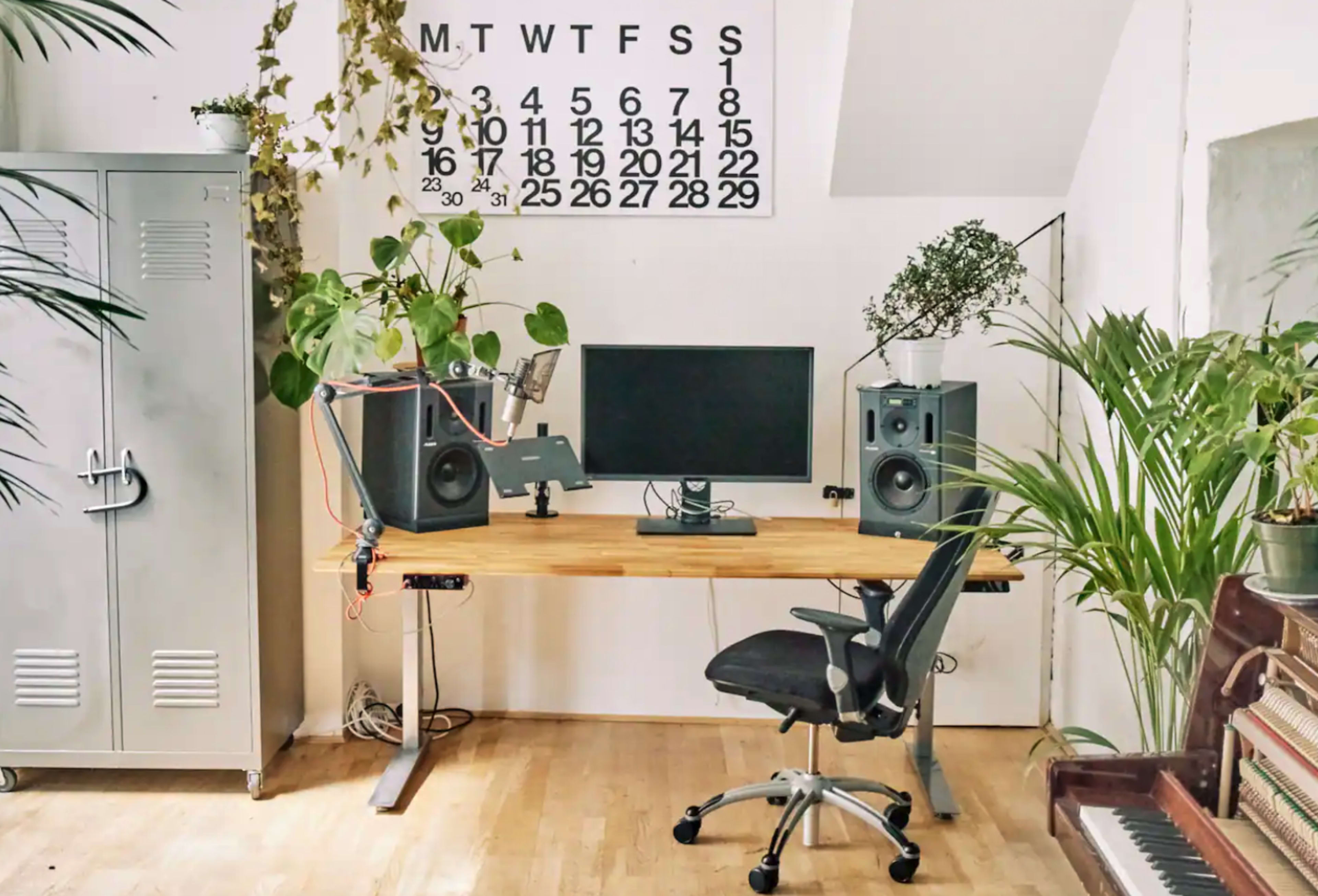 A tidy workspace featuring a wooden desk with a computer monitor, two speakers, a microphone, a chair, and various indoor plants against a wall with a calendar.