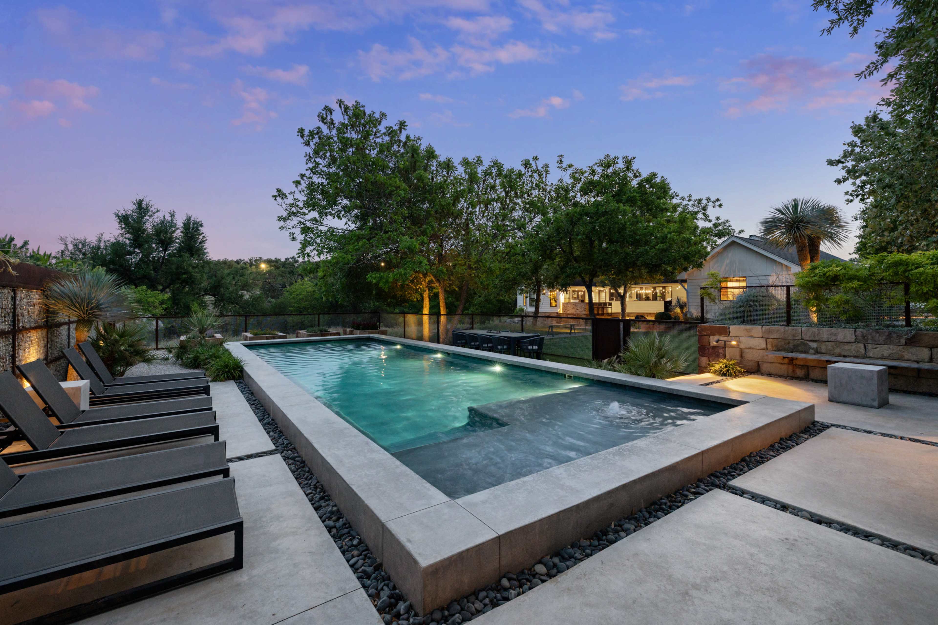 The image shows a modern outdoor pool area surrounded by lounge chairs and decorative landscaping at dusk.