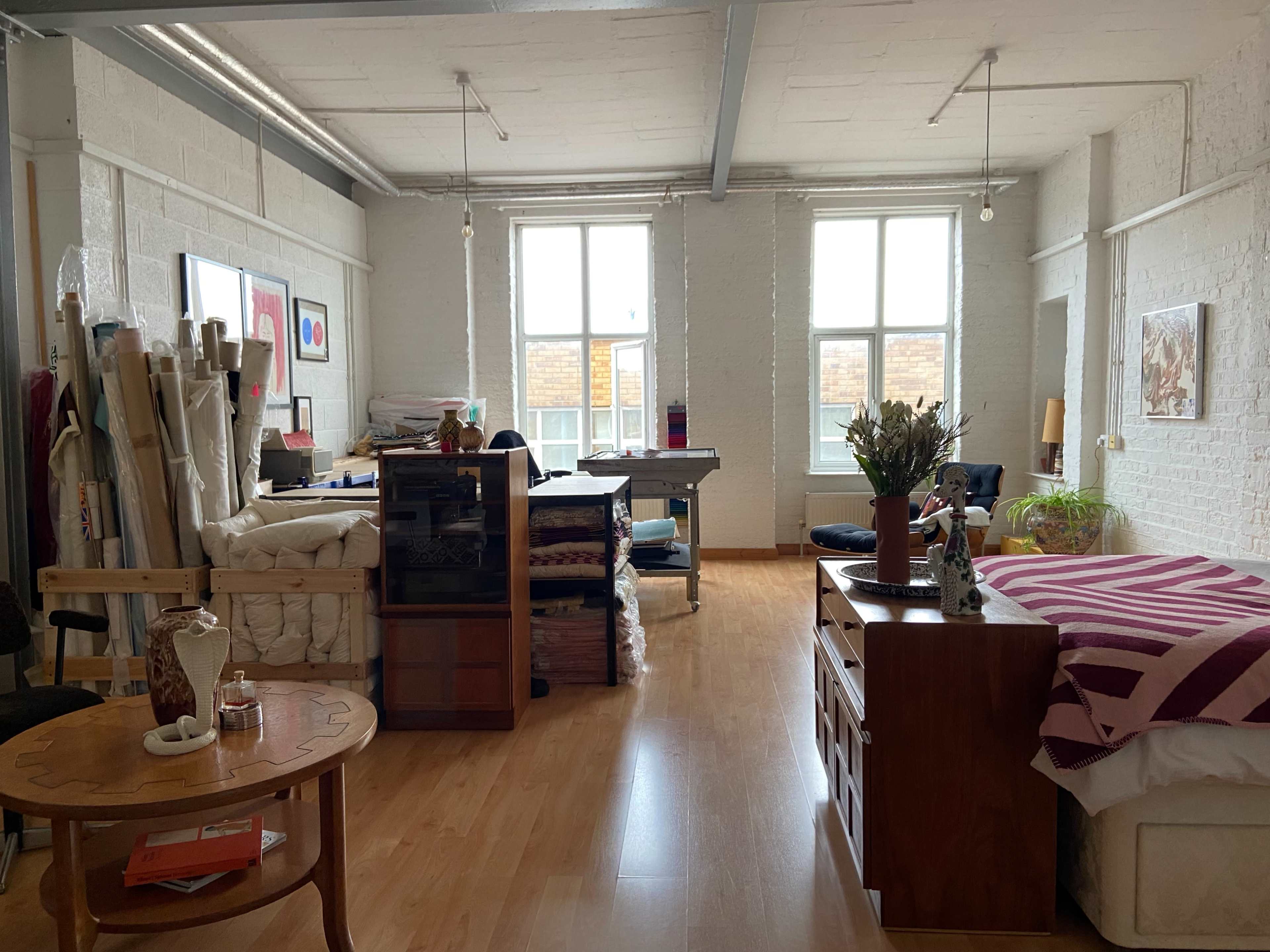 A spacious studio apartment with wooden flooring, large windows, a bed in one corner, and various furniture pieces and art supplies scattered throughout.