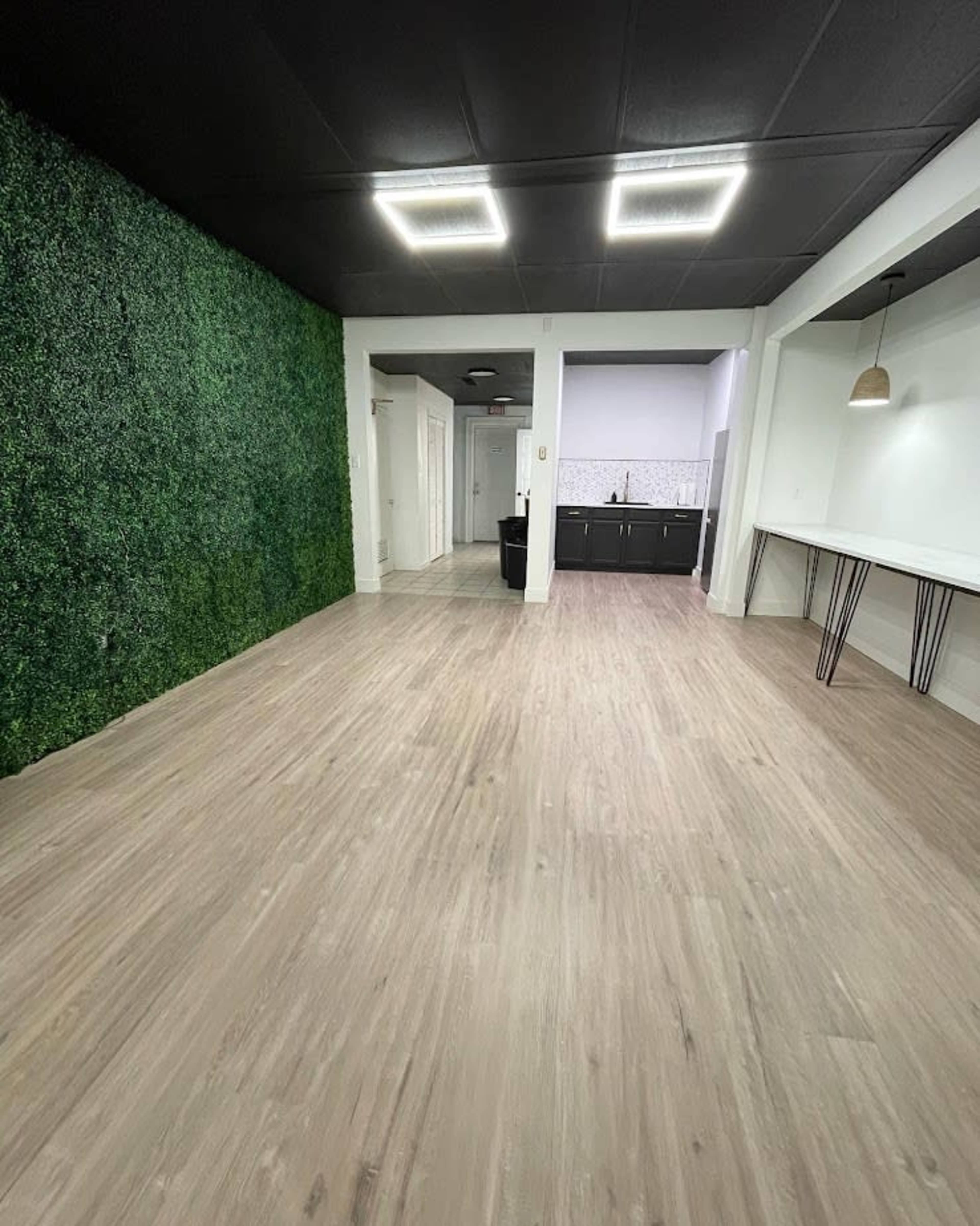 A spacious room with a green plant wall on one side, a kitchen area in the back, and light-colored wood flooring throughout.