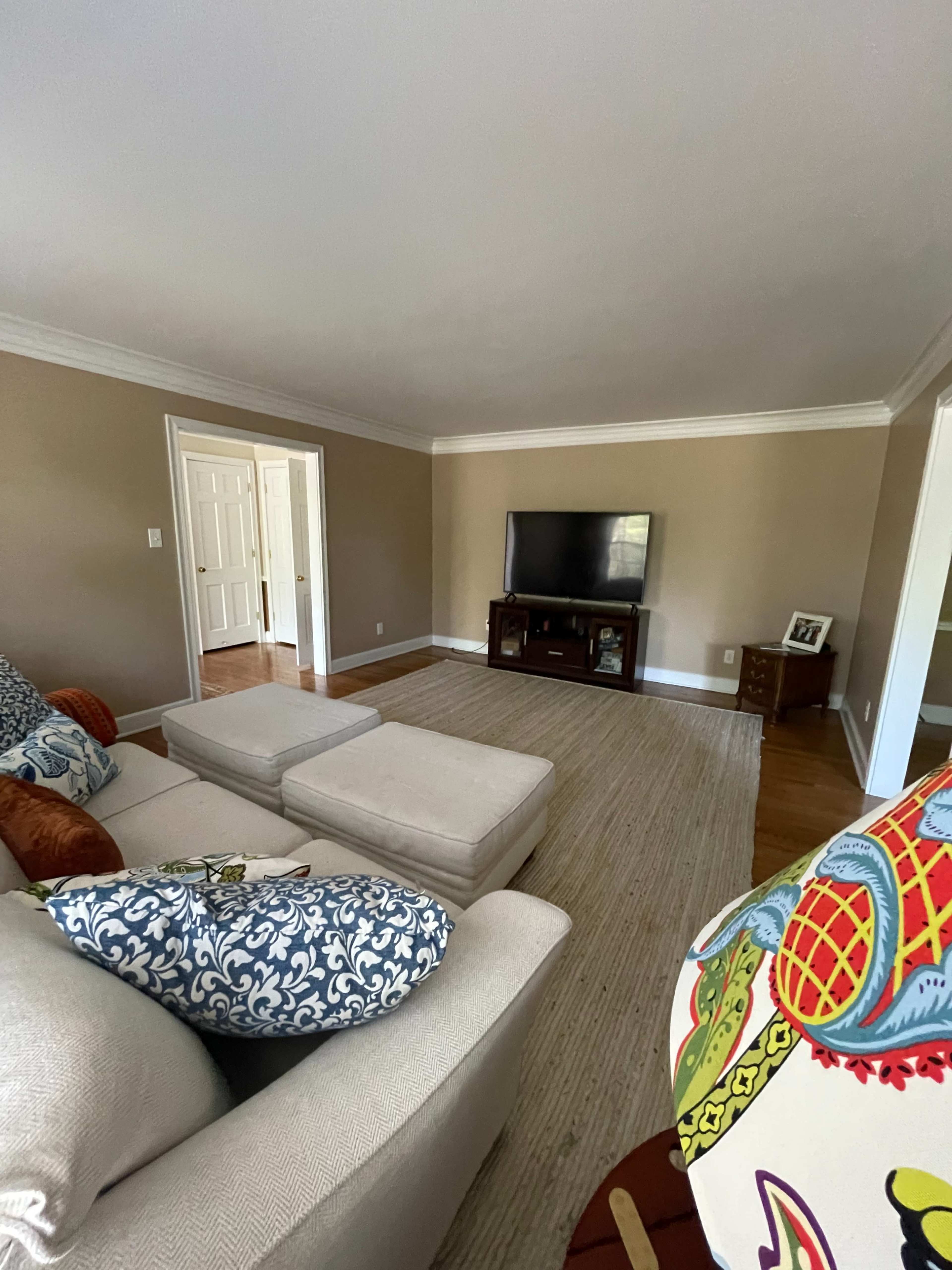 A spacious living room features a sofa, two ottomans, and a television on a low cabinet, with light-colored walls and hardwood floors.