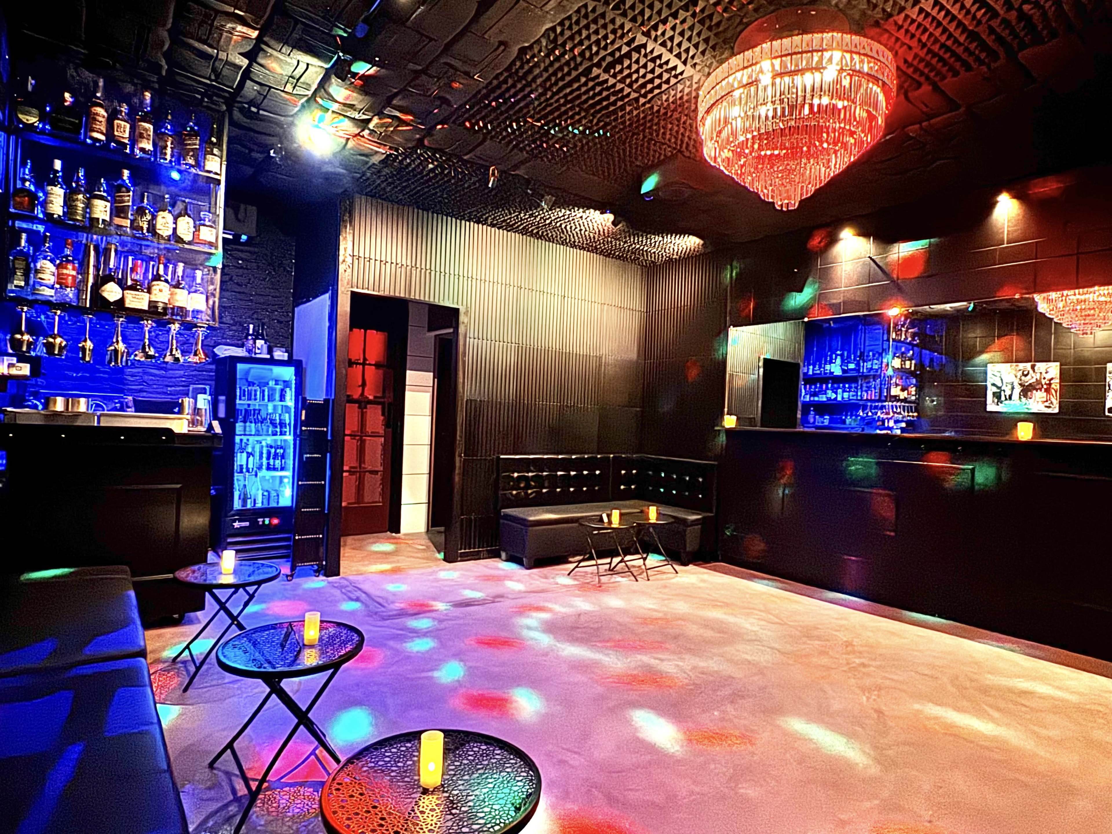 The scene shows a dimly lit bar area with a black interior, shelves filled with bottles, a crystal chandelier, and colorful lights reflecting off the walls.