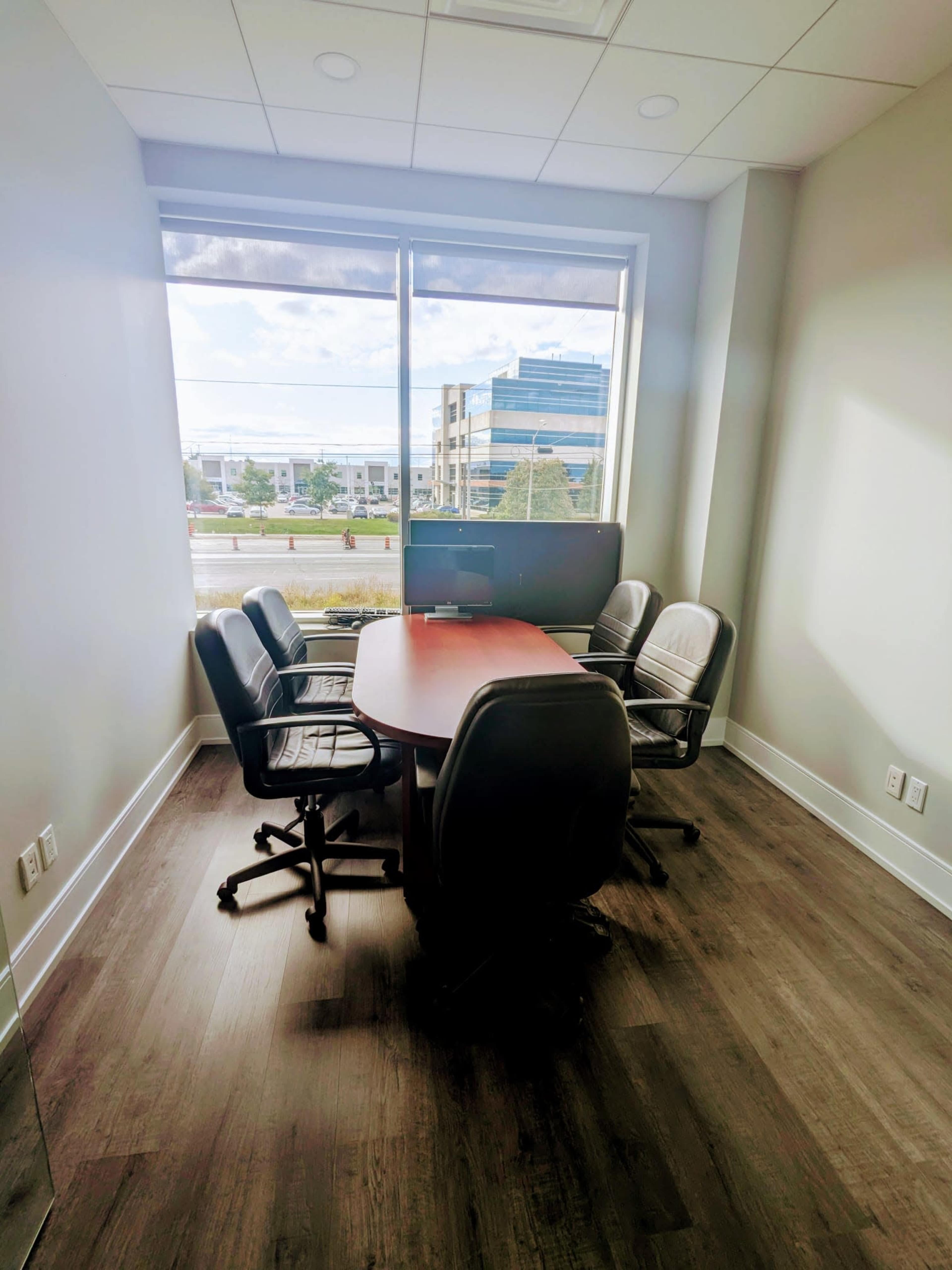 A small conference room with a round table and four black chairs near a large window, overlooking a street and buildings outside.